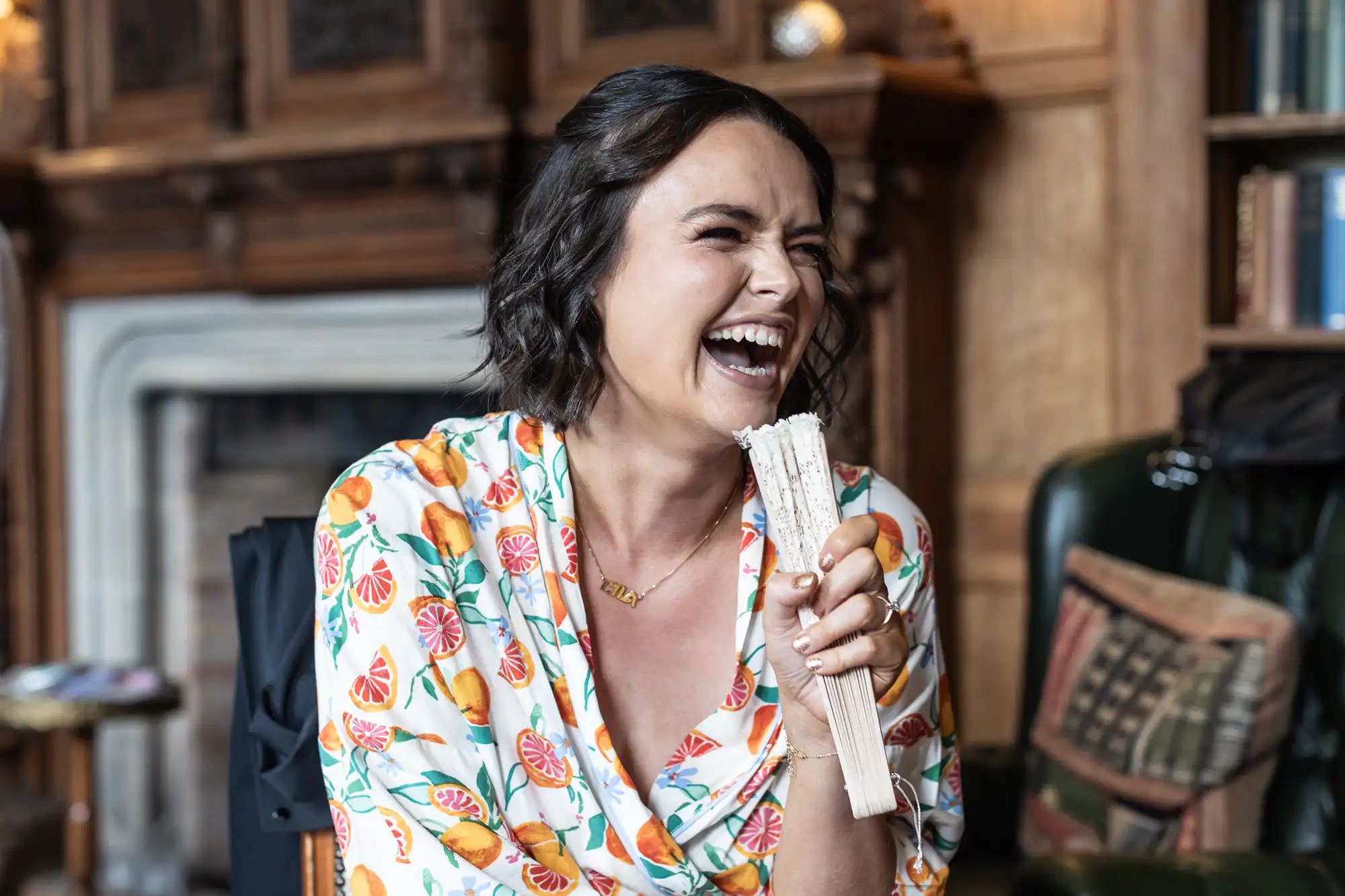 A woman in a fruit-patterned shirt laughs while holding a folding fan, sitting in a room with wooden decor and a fireplace in the background.