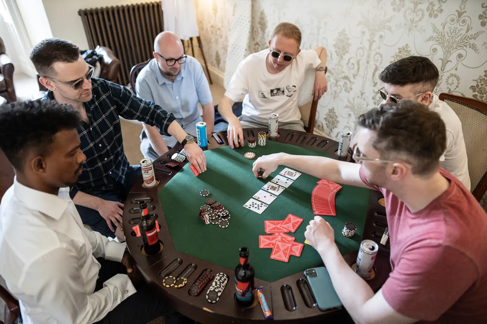 Six men playing poker around a green table, with cards and poker chips visible. Drinks and snacks are scattered on the table.