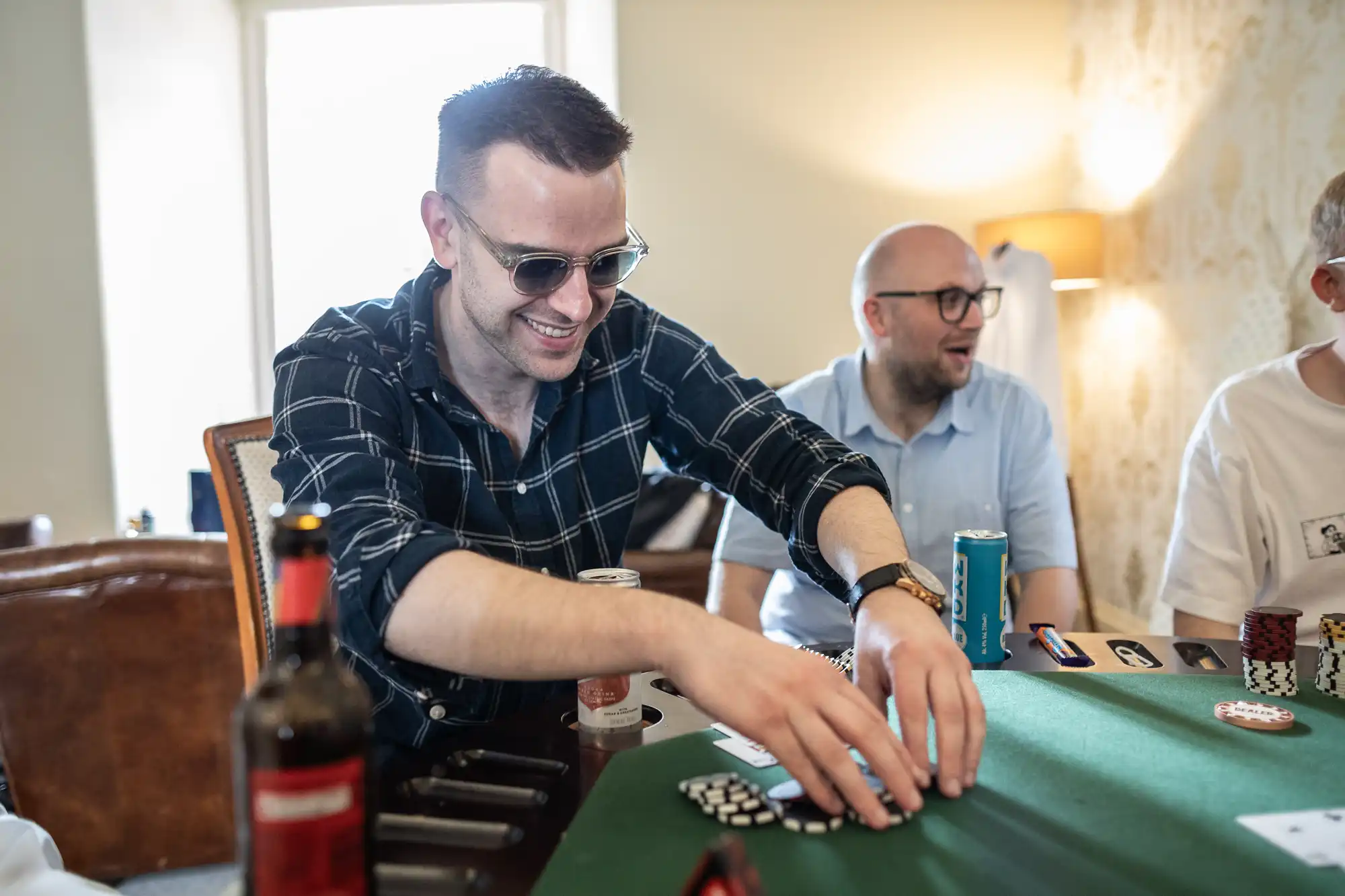 Person wearing sunglasses and a checkered shirt plays poker, smiling as they gather chips. Another person wearing glasses is seated next to them. Table has cards, chips, and a drink can.