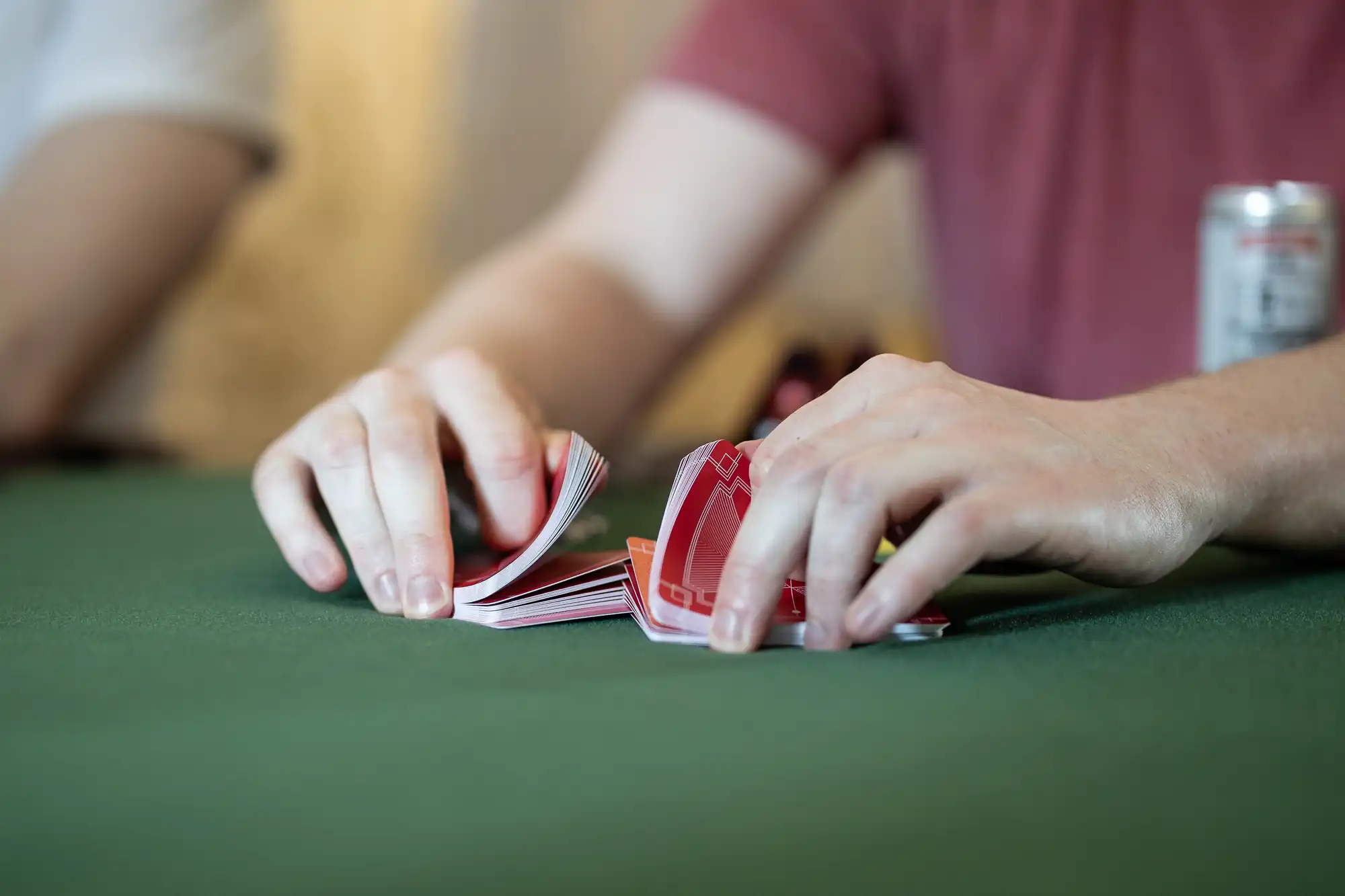 Hands shuffling a deck of playing cards on a green table. A can is visible in the background.