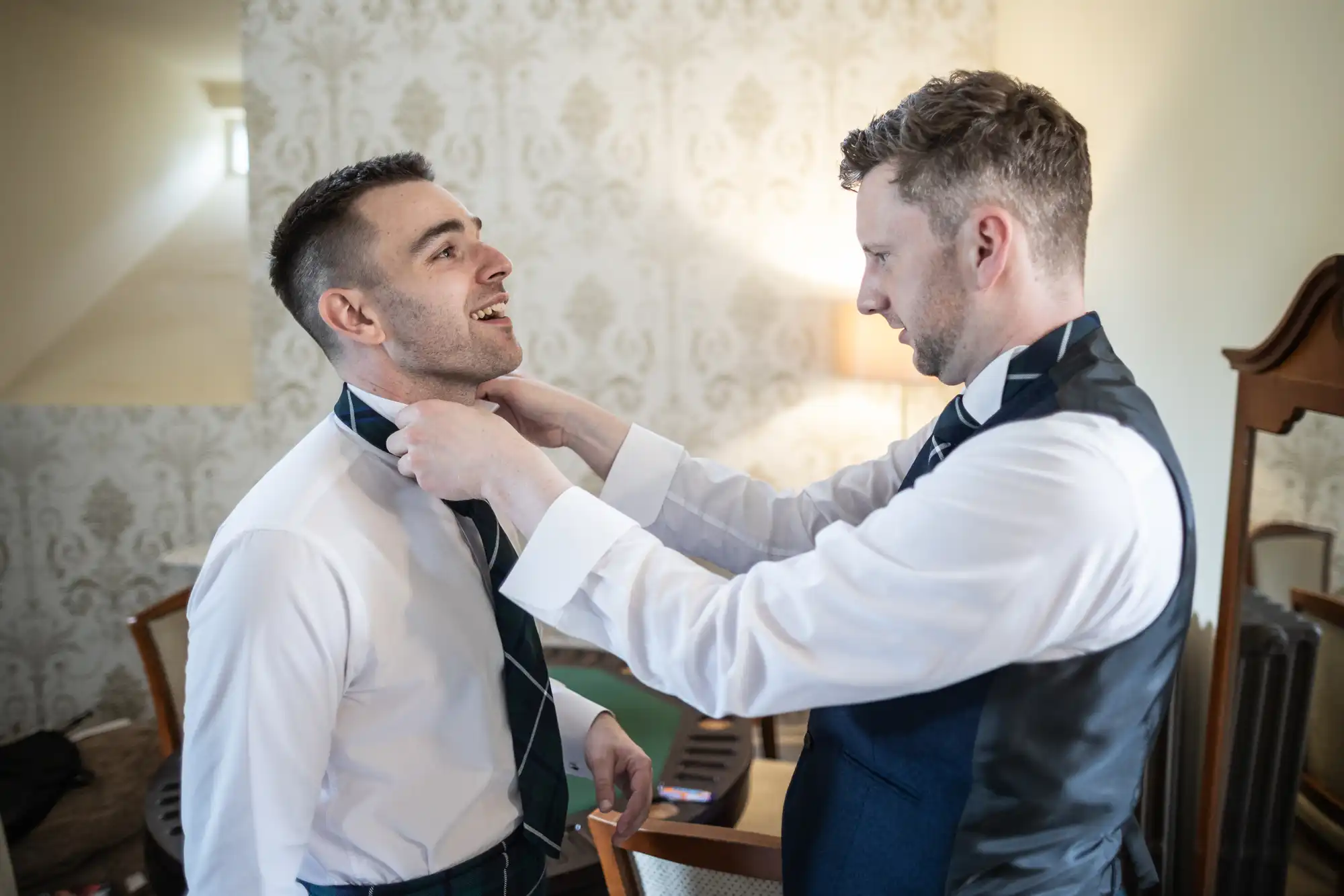 A man adjusts another man's tie in a room with ornate wallpaper, a lamp, and a wooden mirror. Both are dressed in formal attire.