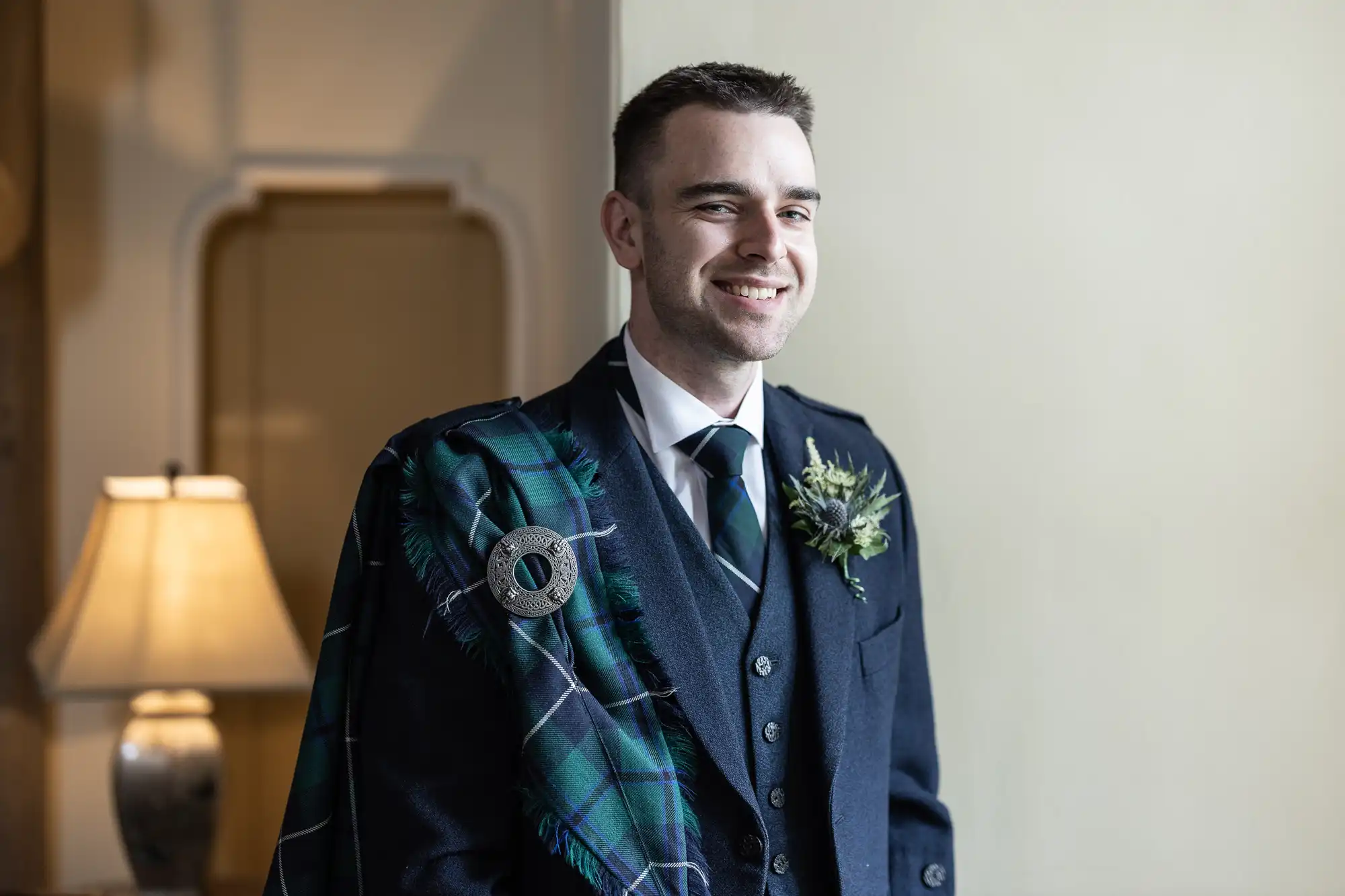 A man in a formal suit and tartan sash stands indoors with a boutonniere on his lapel, smiling. A lamp and a wall are in the background.