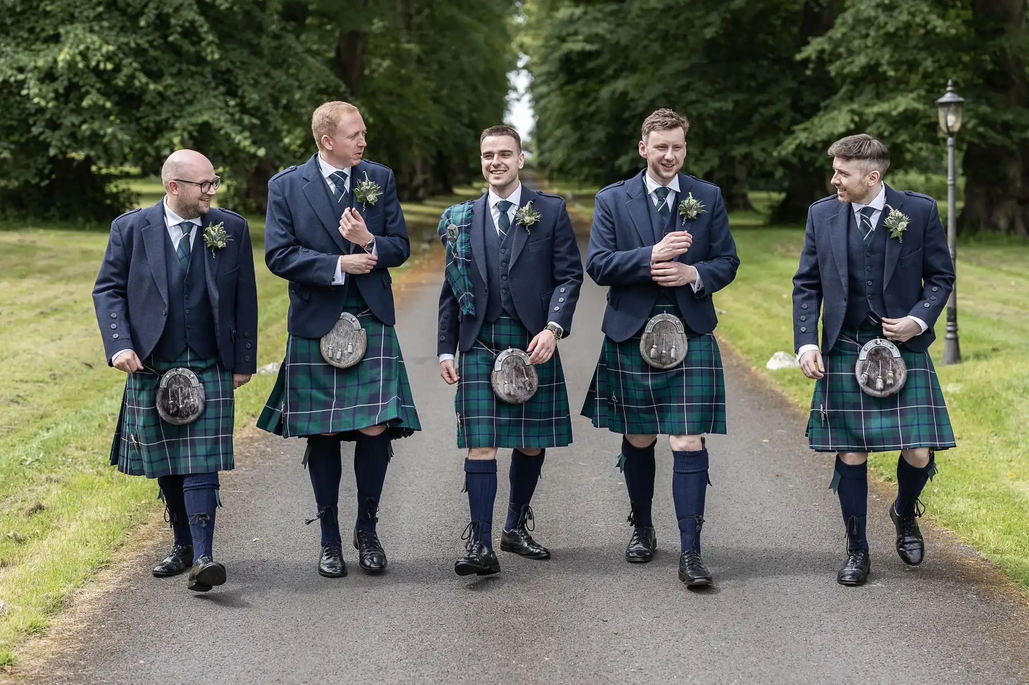 Five men in matching kilts and jackets walk side by side on a tree-lined path, smiling and conversing.