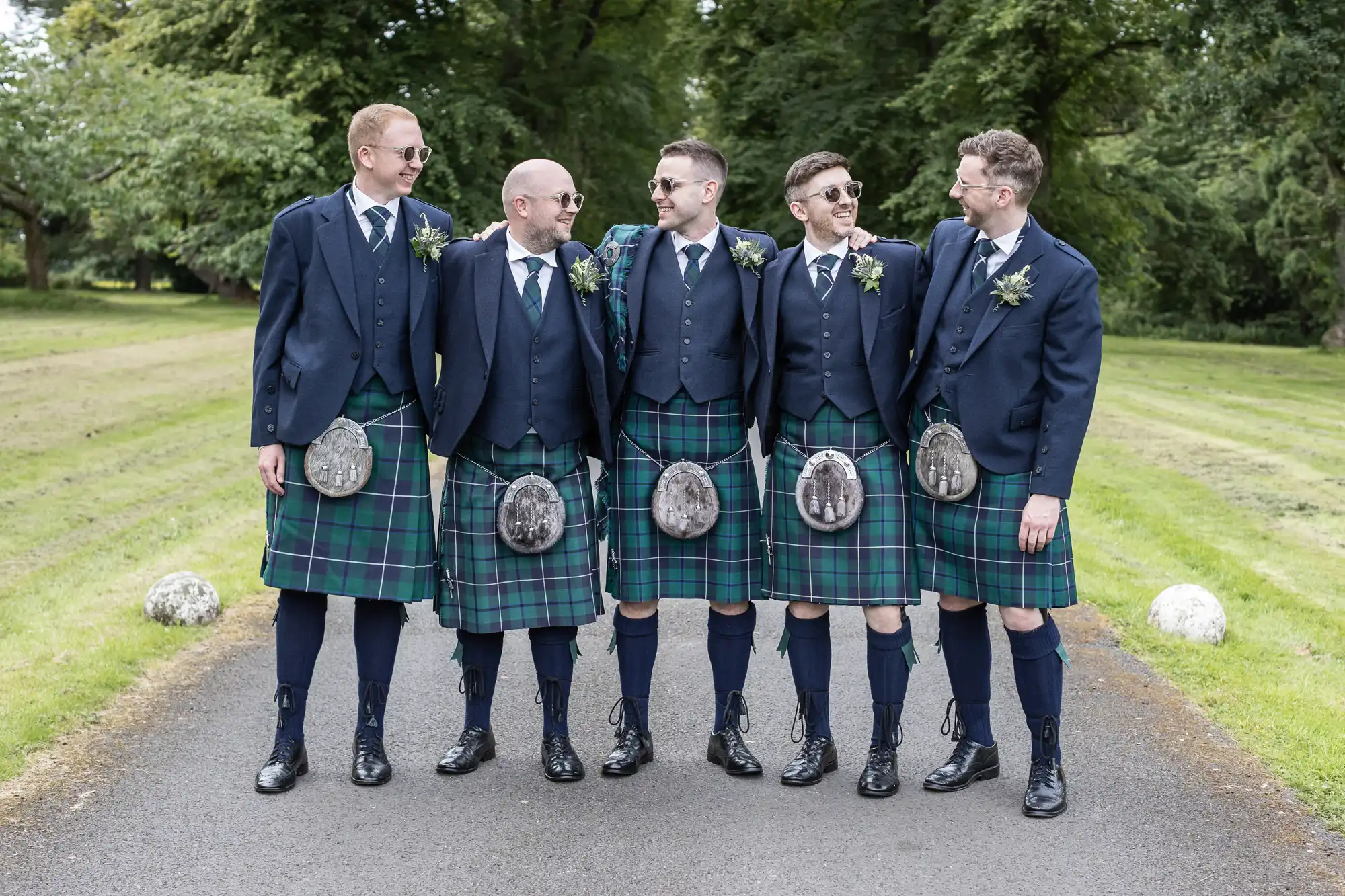 Five men in sunglasses wearing matching blue jackets and green tartan kilts stand together on a path, surrounded by greenery.