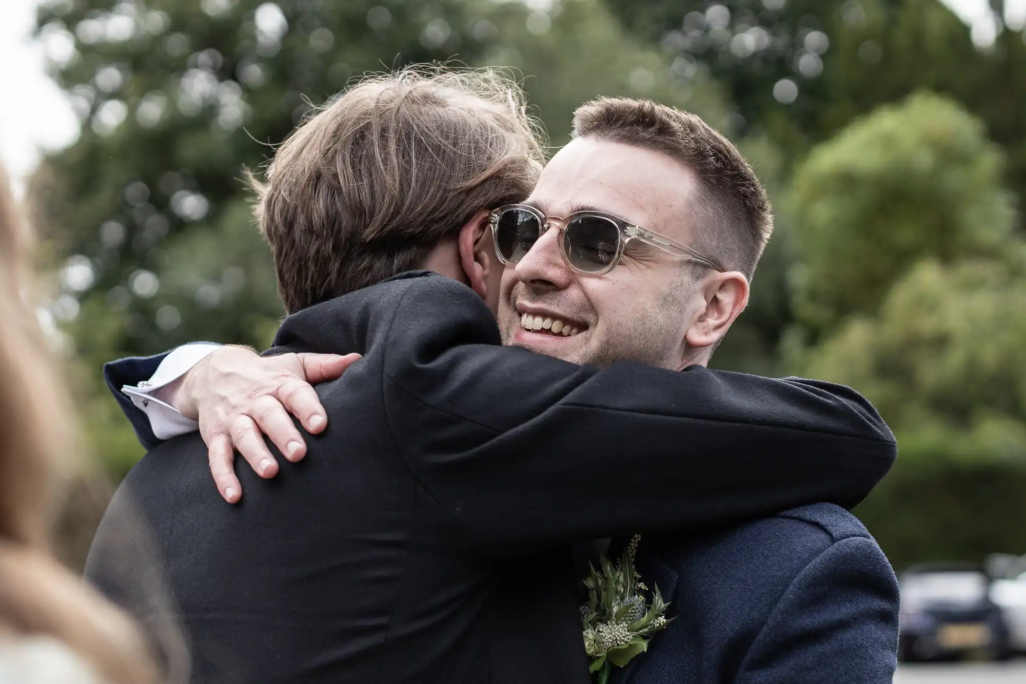 Two men in suits hugging outdoors; the man facing the camera is wearing sunglasses and smiling.