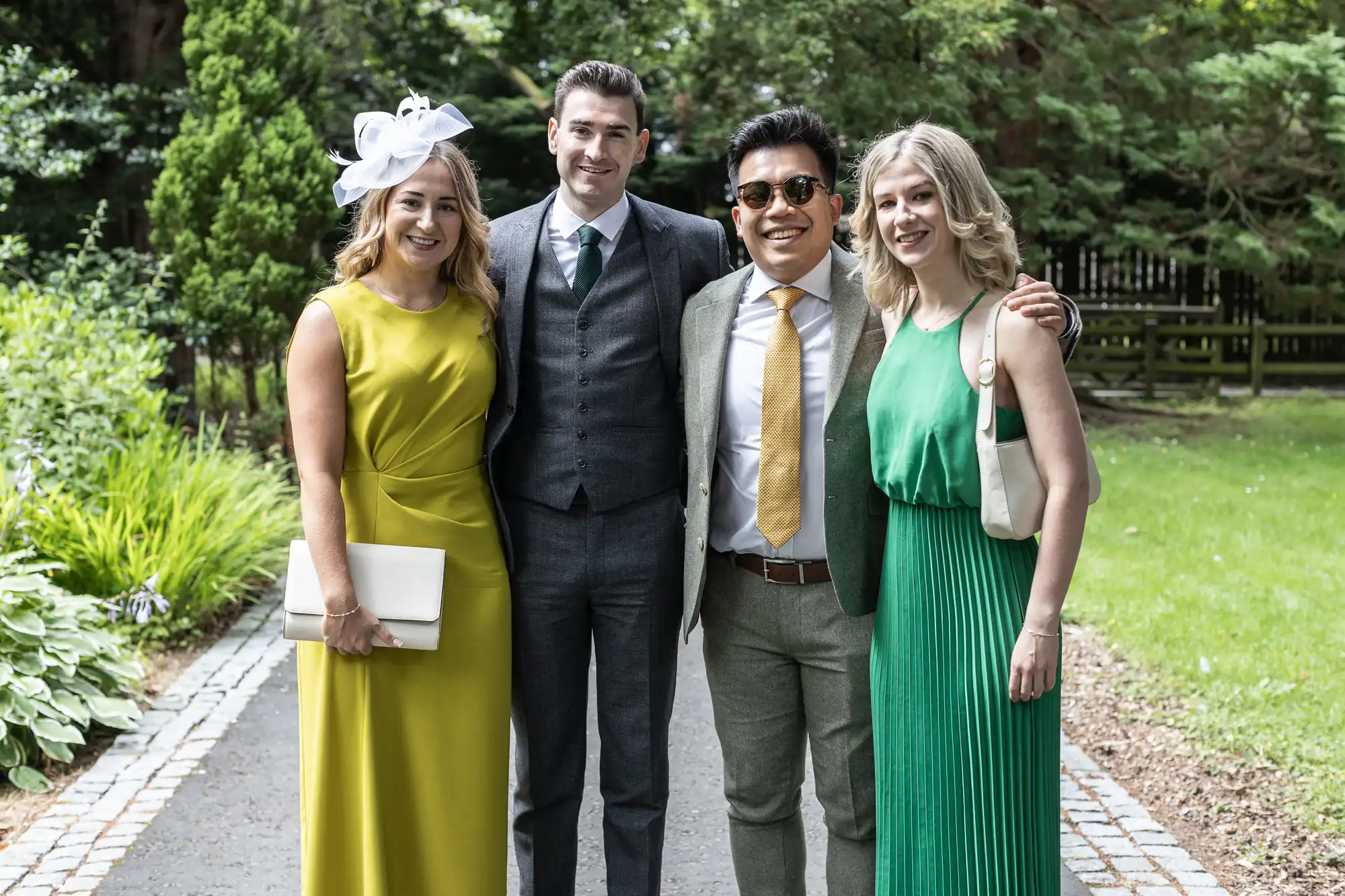 Four people in formal attire standing on a garden path, smiling at the camera. The group includes two men in suits and two women in colorful dresses, one with a white hat.