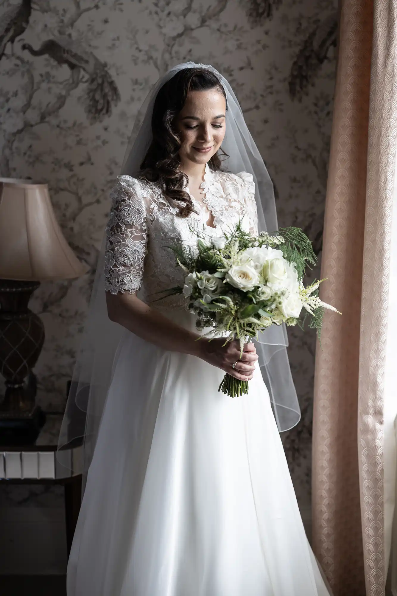 Bride in a white gown and veil holds a bouquet of white flowers, standing near a window with light filtering in, with floral wallpaper and a lamp in the background.