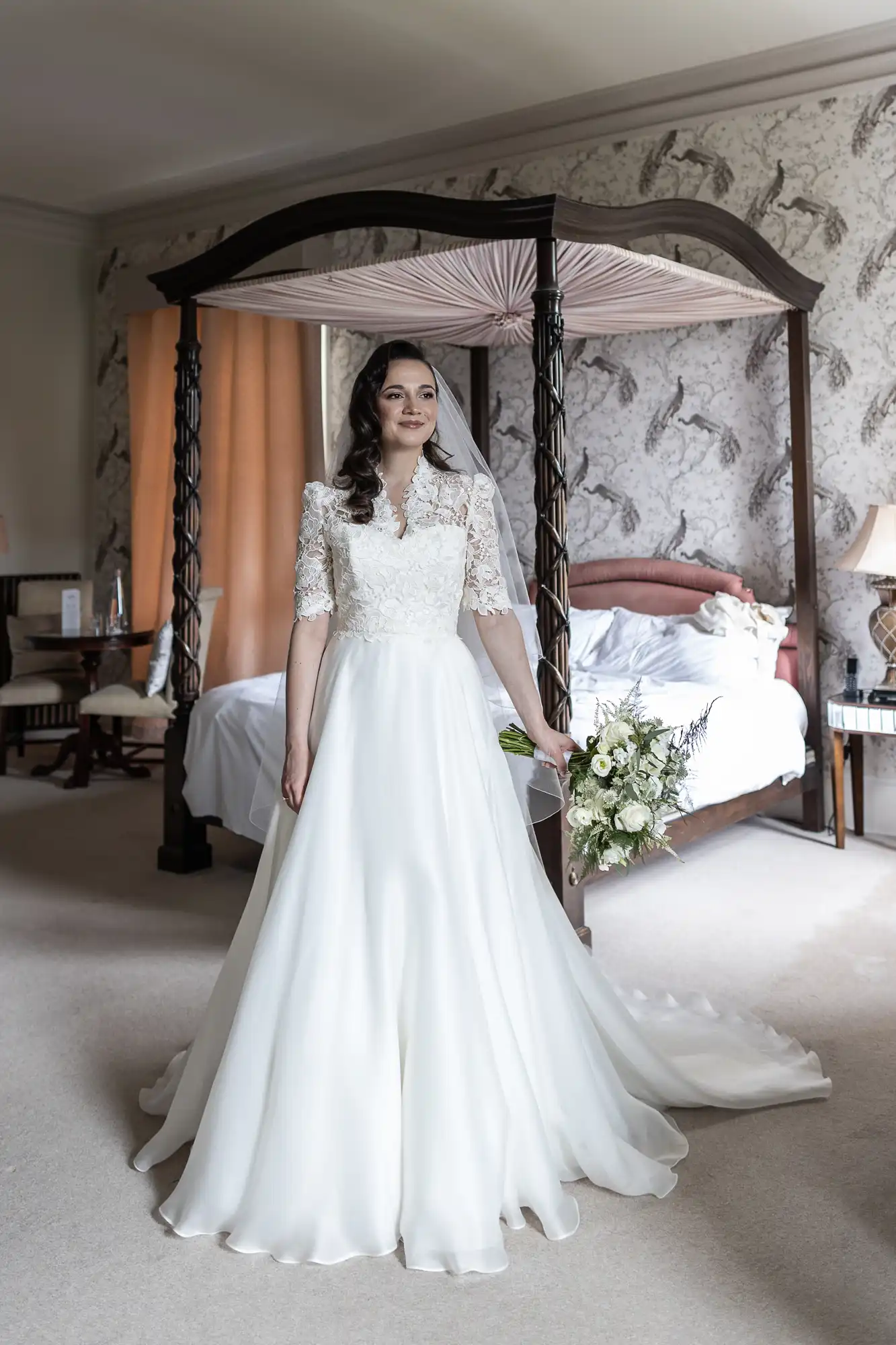A bride in a white gown stands in a bedroom with floral wallpaper. She holds a bouquet and wears a veil, with an elegant bed and curtains in the background.