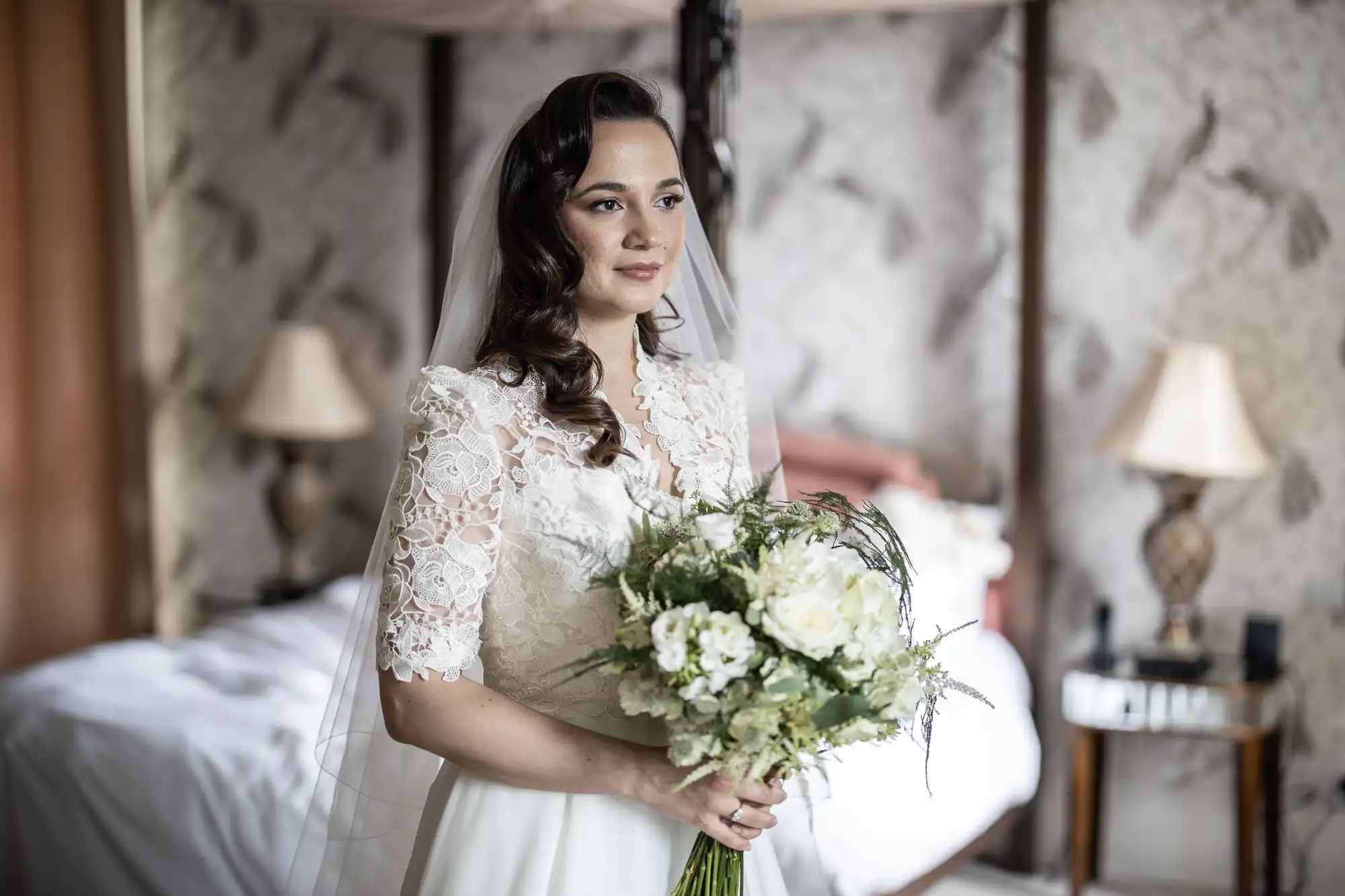 Bride in a lace wedding dress holding a bouquet of white flowers, standing in a room with patterned wallpaper and a bed in the background.