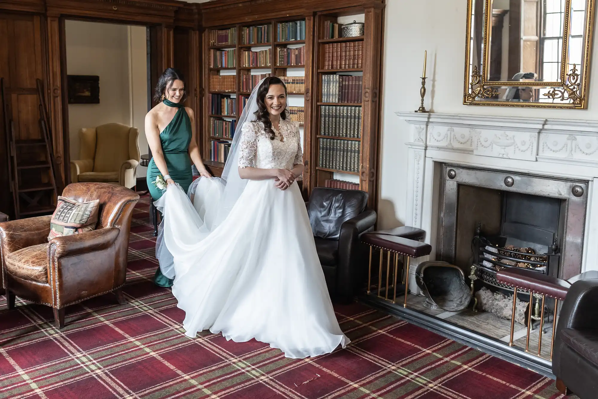 A bride in a white gown stands in a room with bookshelves and a fireplace. A person in a green dress assists with her train.