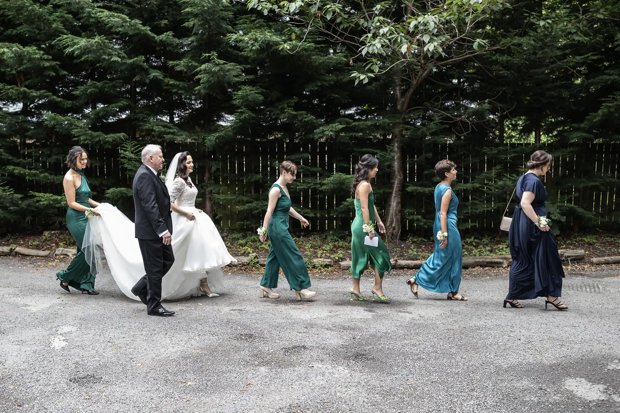 A bride in a white dress walks with bridesmaids in green and blue dresses, accompanied by an older man in a suit, along a tree-lined path.