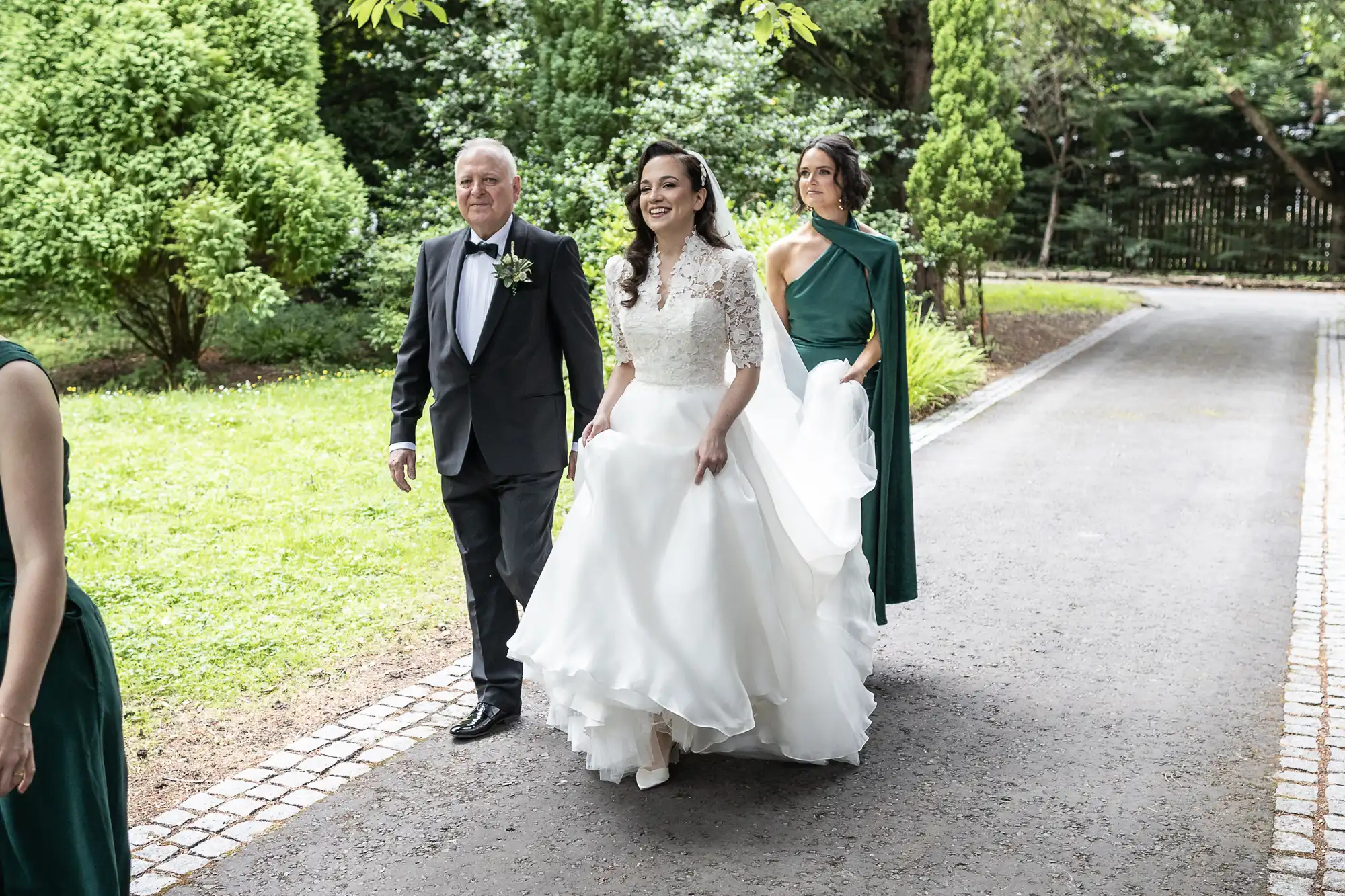 A bride in a white dress walks on a path alongside a man in a suit and a woman in a green dress, set against a backdrop of greenery.