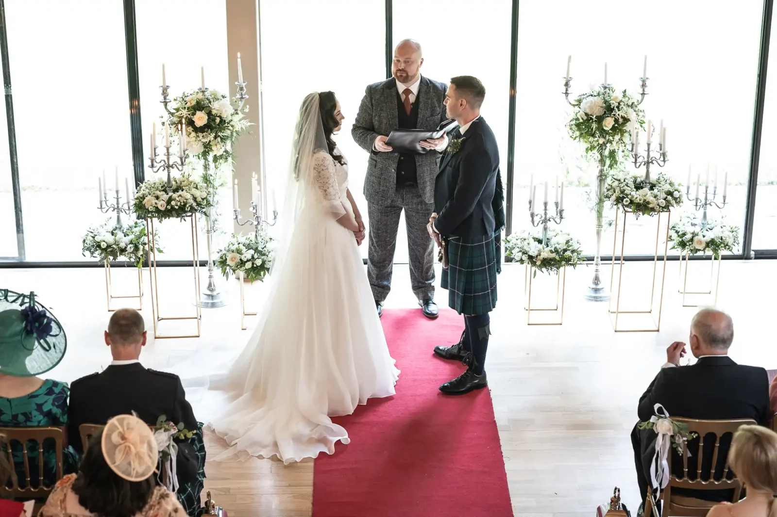 A couple is getting married indoors at The Chapel at Carberry Tower, on a red carpet, with a person officiating the ceremony. Guests are seated, and decorative flowers and candelabras are in the background.