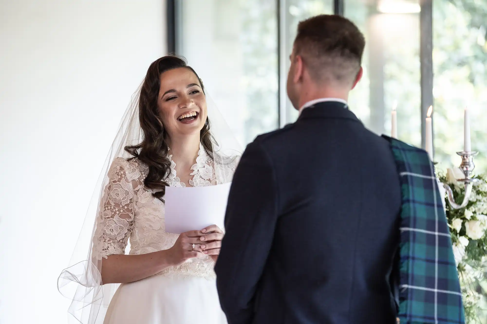 A bride in a lace dress reads vows to a groom wearing a kilt and jacket. They are indoors with a window and flowers in the background.