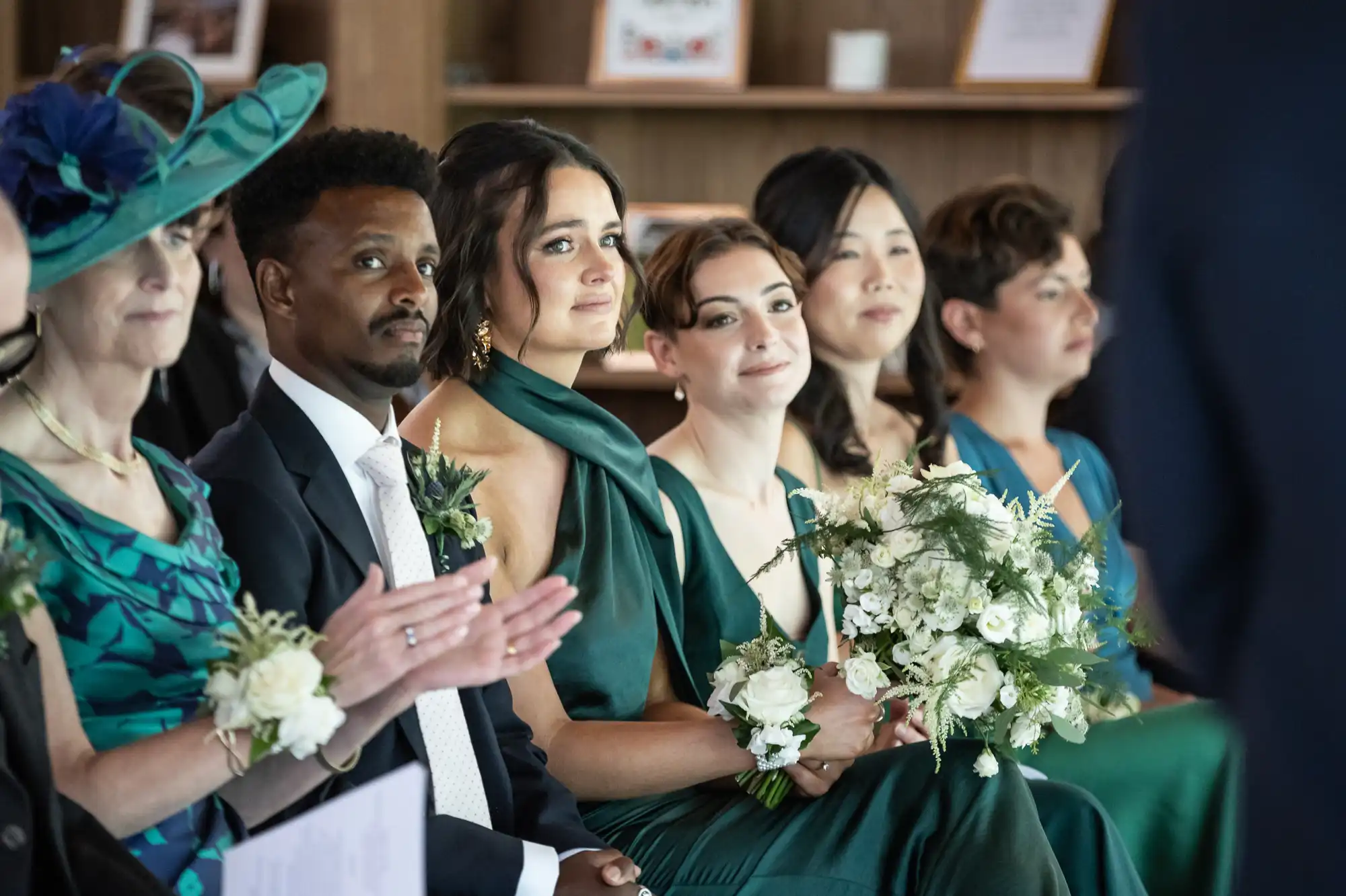 A group of people in formal attire sit together, some holding flower bouquets.