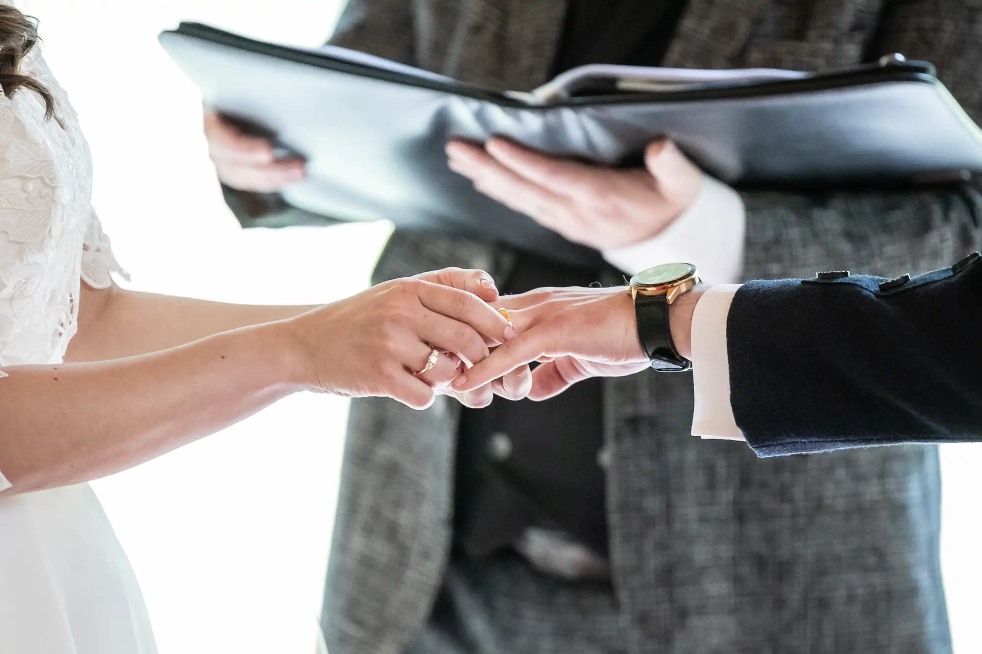 Couple exchanging rings during a wedding ceremony, with an officiant holding a book in the background.