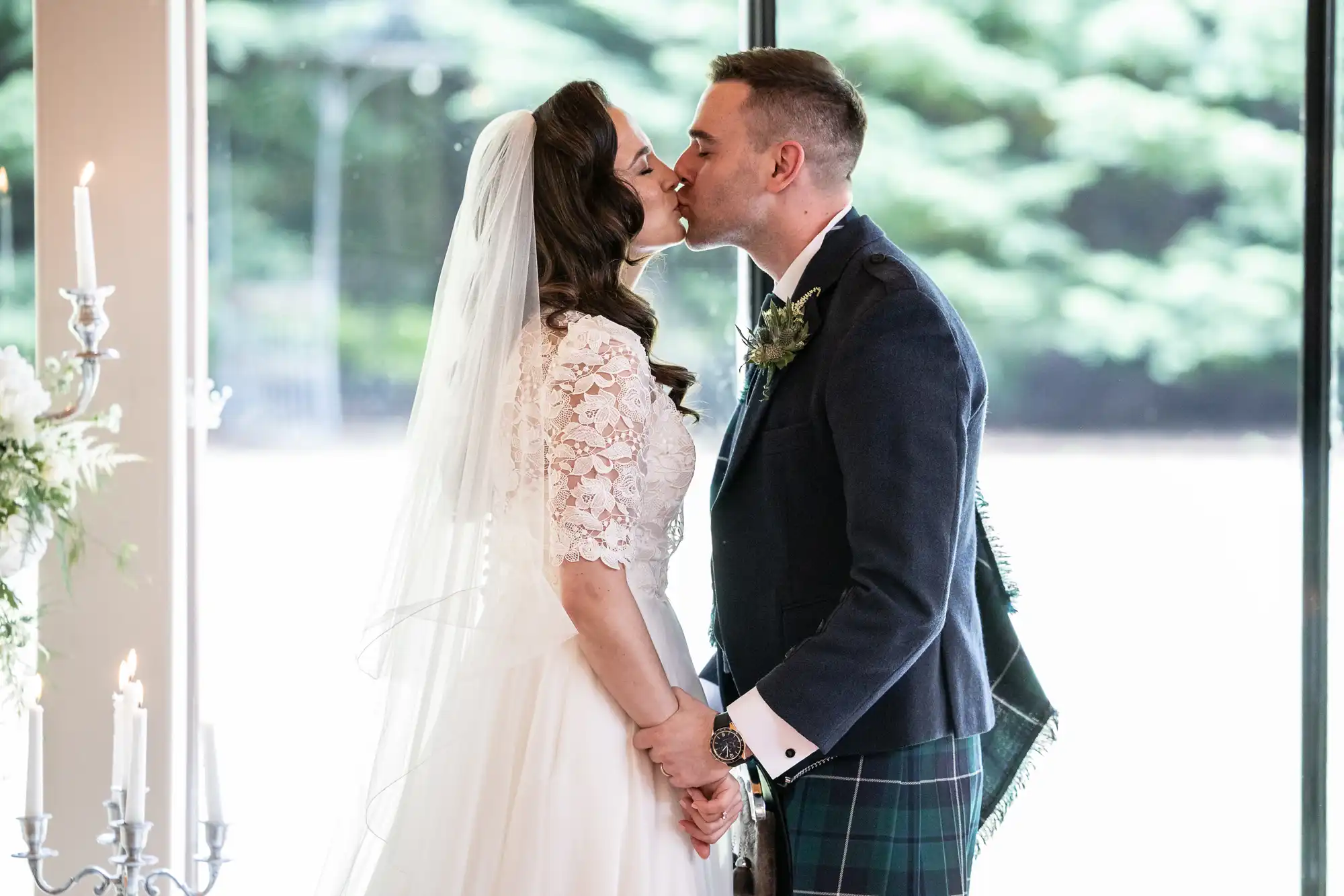 A bride and groom kiss during their wedding ceremony, holding hands. She wears a white dress and veil; he is in a kilt and jacket. They stand in front of large windows with greenery outside.