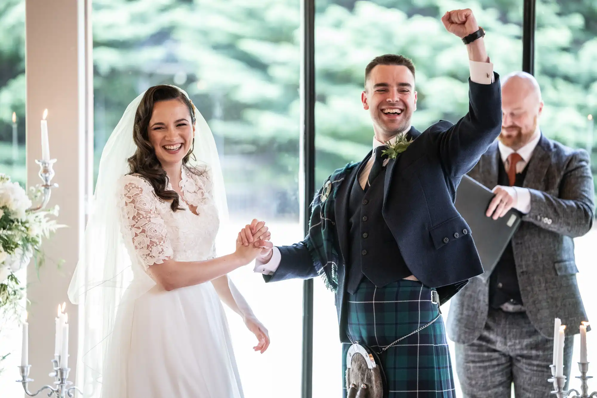Bride and groom holding hands and smiling joyfully during a wedding ceremony, with the groom raising a fist in celebration. An officiant stands nearby.
