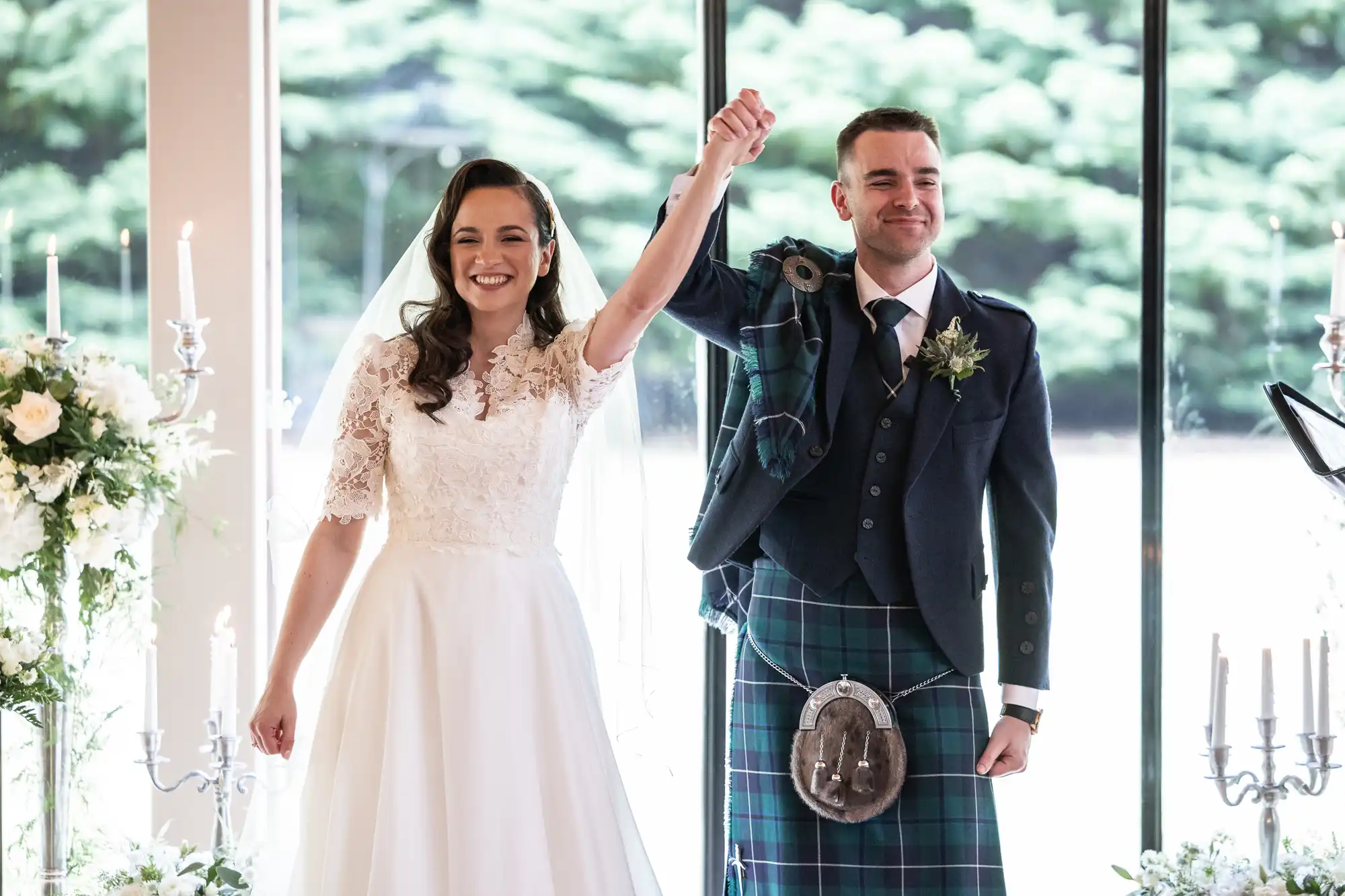 Bride in a white dress and groom in a kilt celebrate joyfully indoors, holding hands aloft, with floral decorations and large windows in the background.