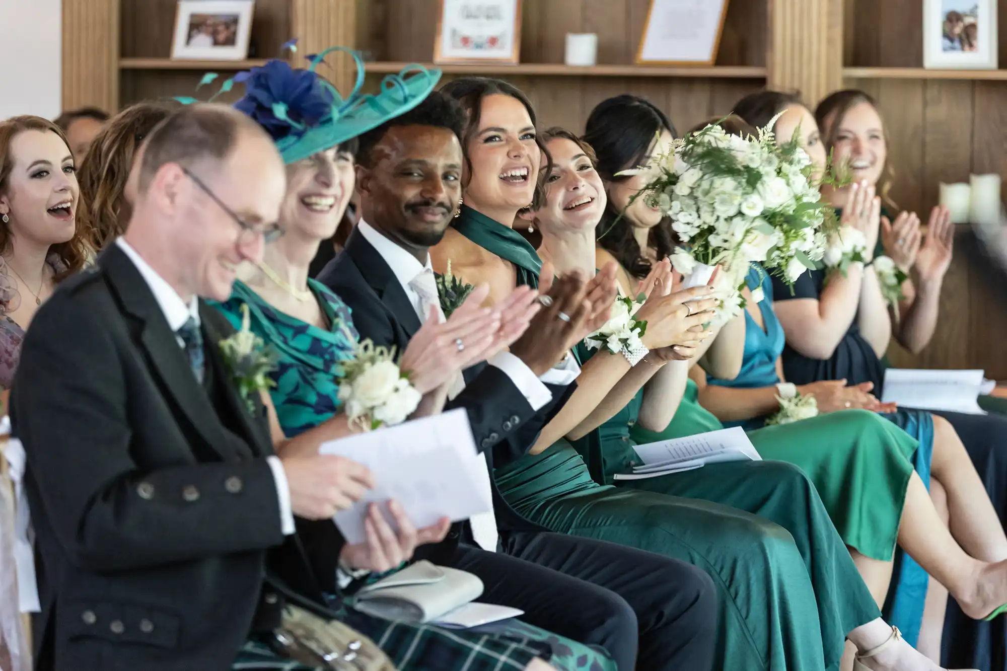 A group of smiling and clapping people sit in a row at a formal event, wearing formal attire and holding bouquets. Books and framed items are on wooden shelves in the background.