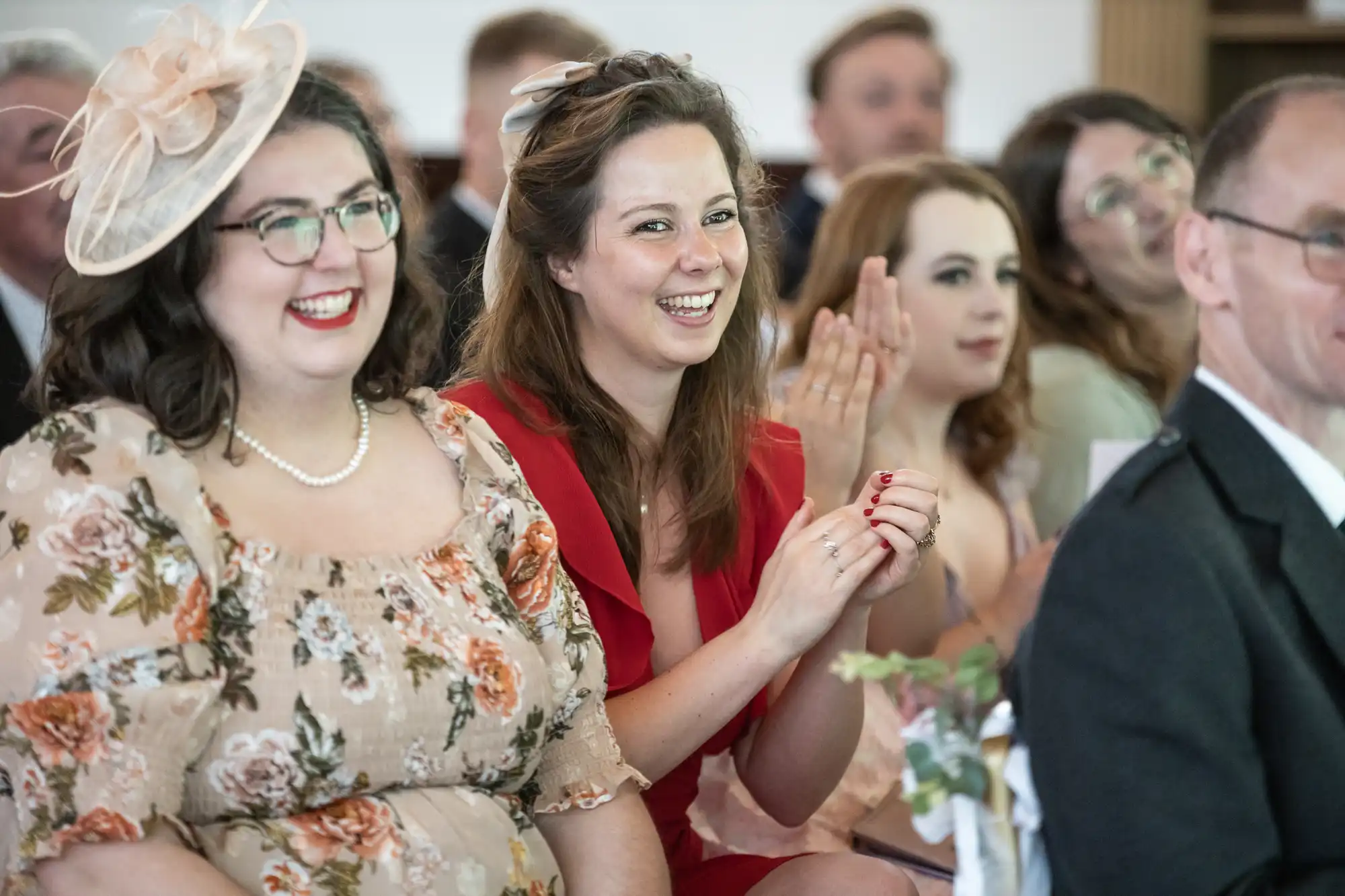 People sitting in an audience, smiling and clapping. Two women in the foreground are wearing floral and red dresses.