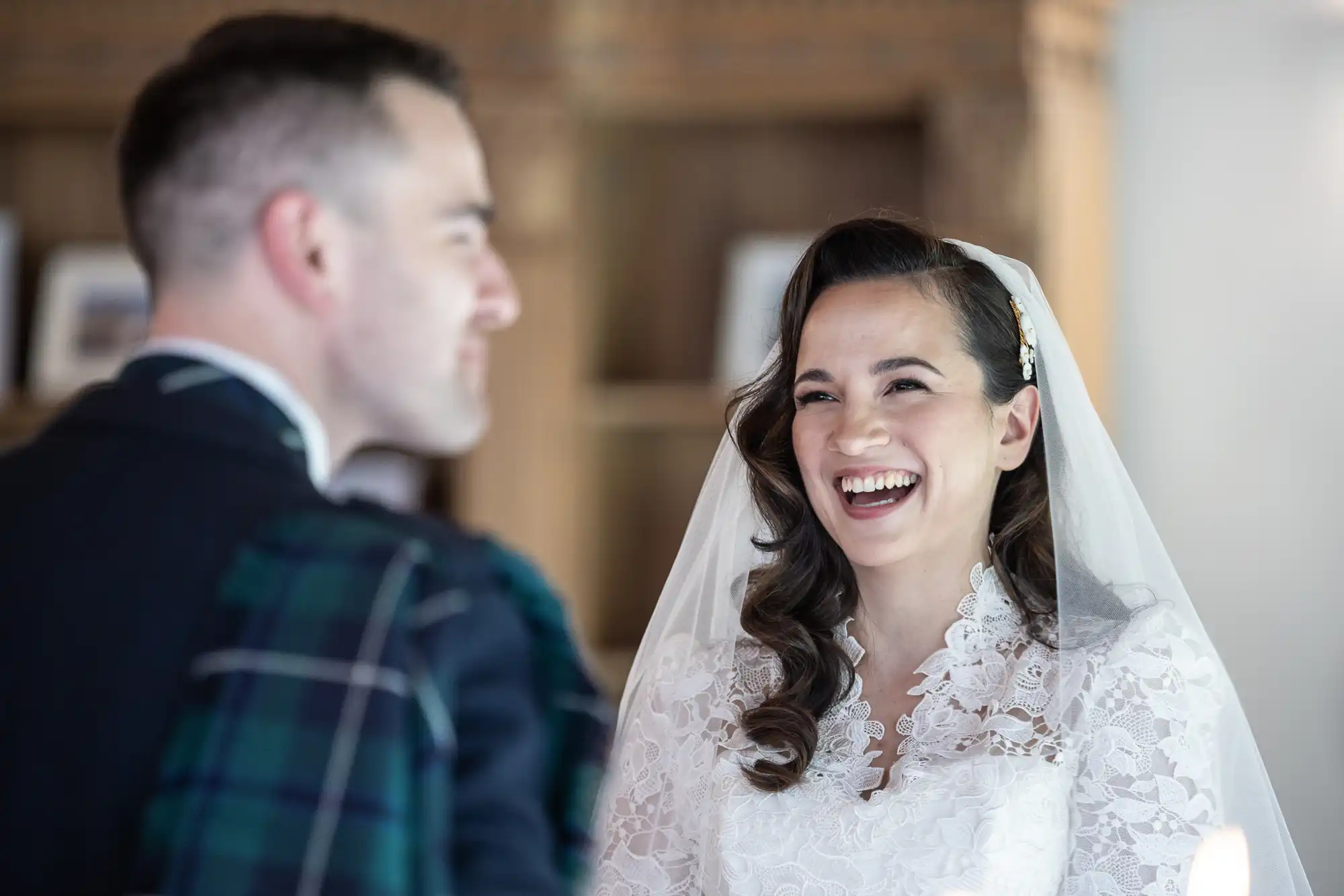 A bride in a lace dress and veil smiles while looking at a groom in a dark outfit and tartan sash. They are indoors with wooden shelves in the background.