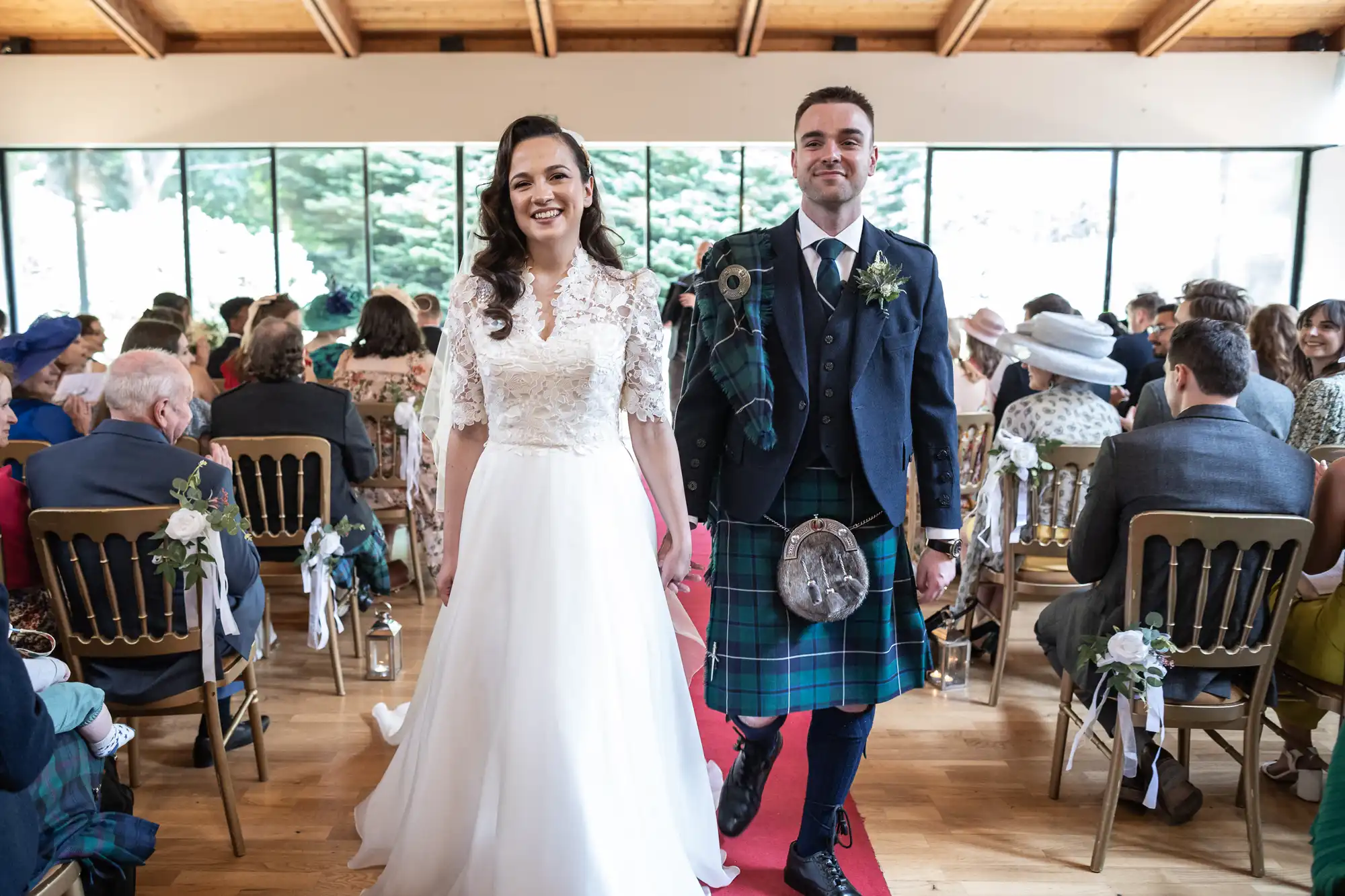 A bride in a white dress and a groom in a kilt walk down the aisle smiling, with guests seated on either side in a bright venue.