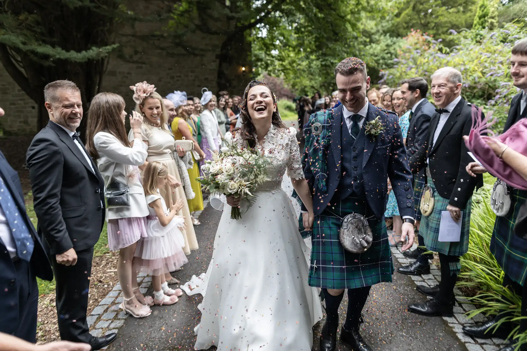 A couple in wedding attire walks through a cheering crowd outdoors. The bride holds a bouquet and laughs, while the groom, wearing a kilt, smiles. Guests throw confetti.