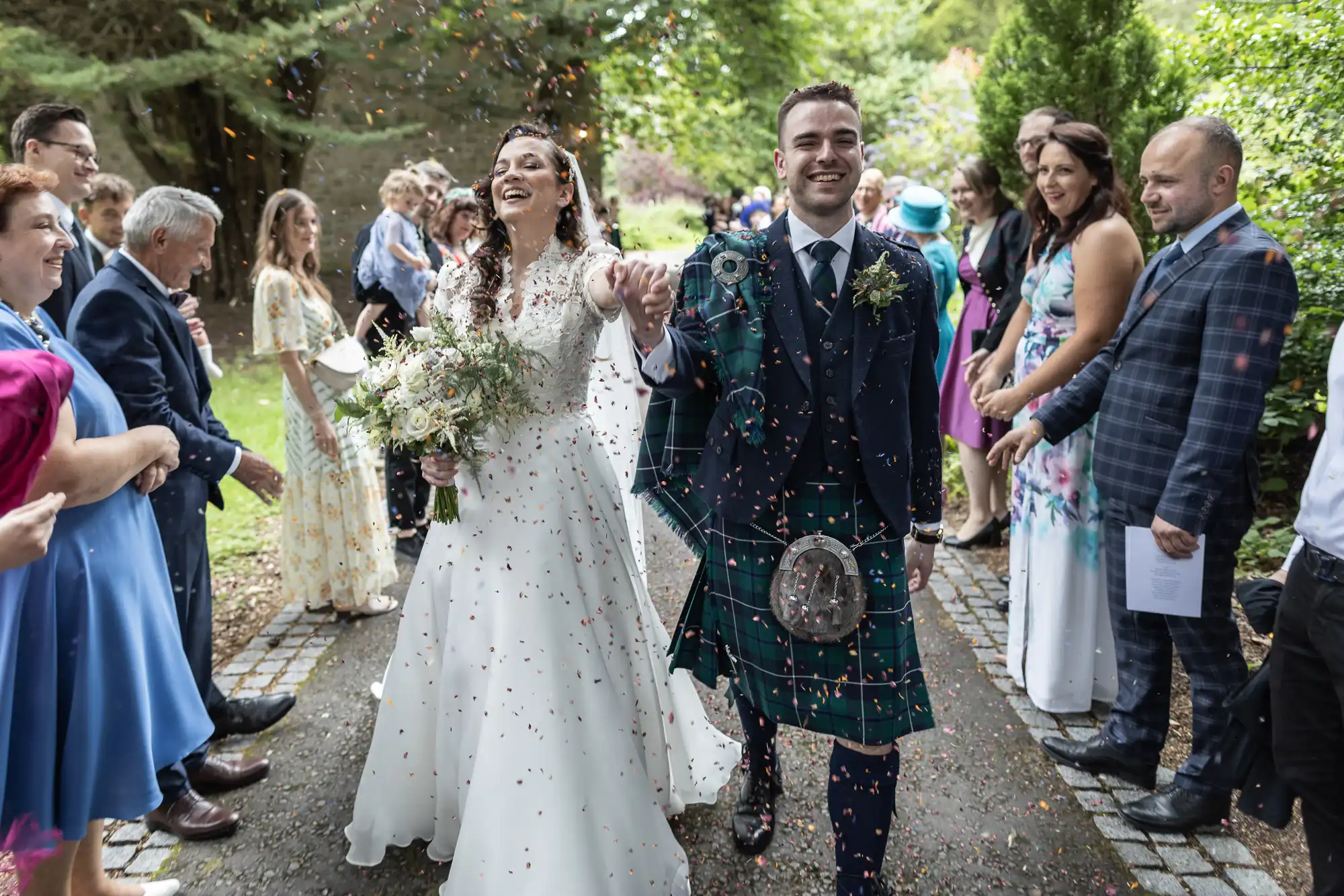A bride and groom walk joyfully down an outdoor path, surrounded by smiling guests who are tossing confetti. The groom wears a kilt, and the bride holds a bouquet.