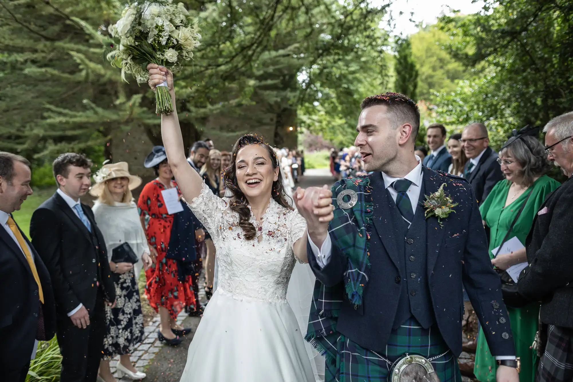 Bride and groom celebrate while walking through a crowd of cheering guests at an outdoor wedding. The bride holds up a bouquet, and the groom wears a kilt.