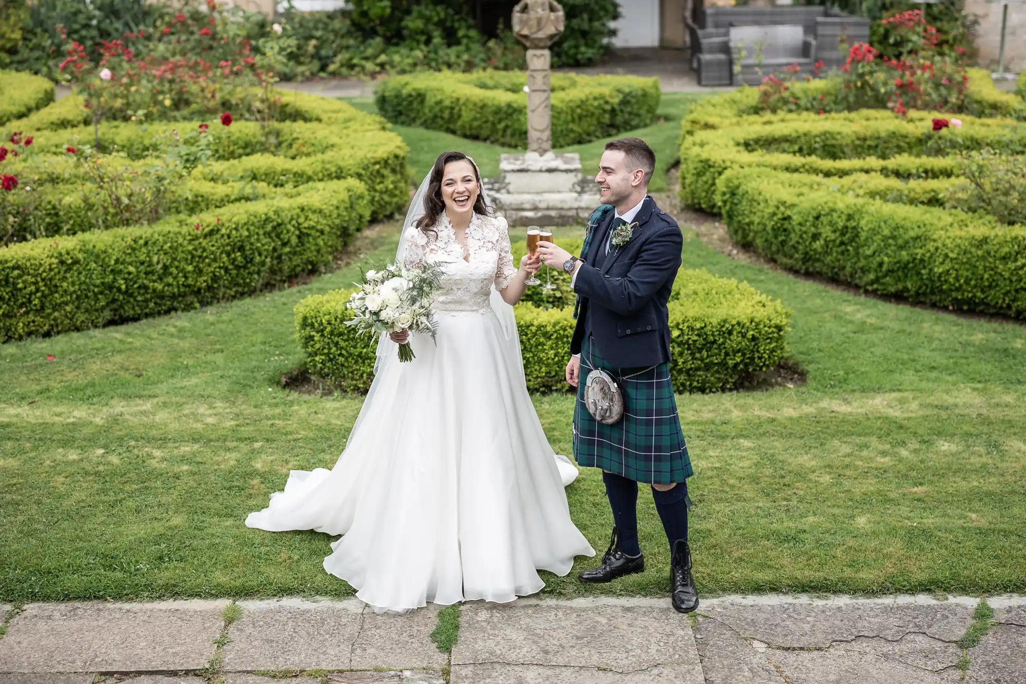 A bride in a white dress and a groom in a kilt toast with drinks in a garden setting.