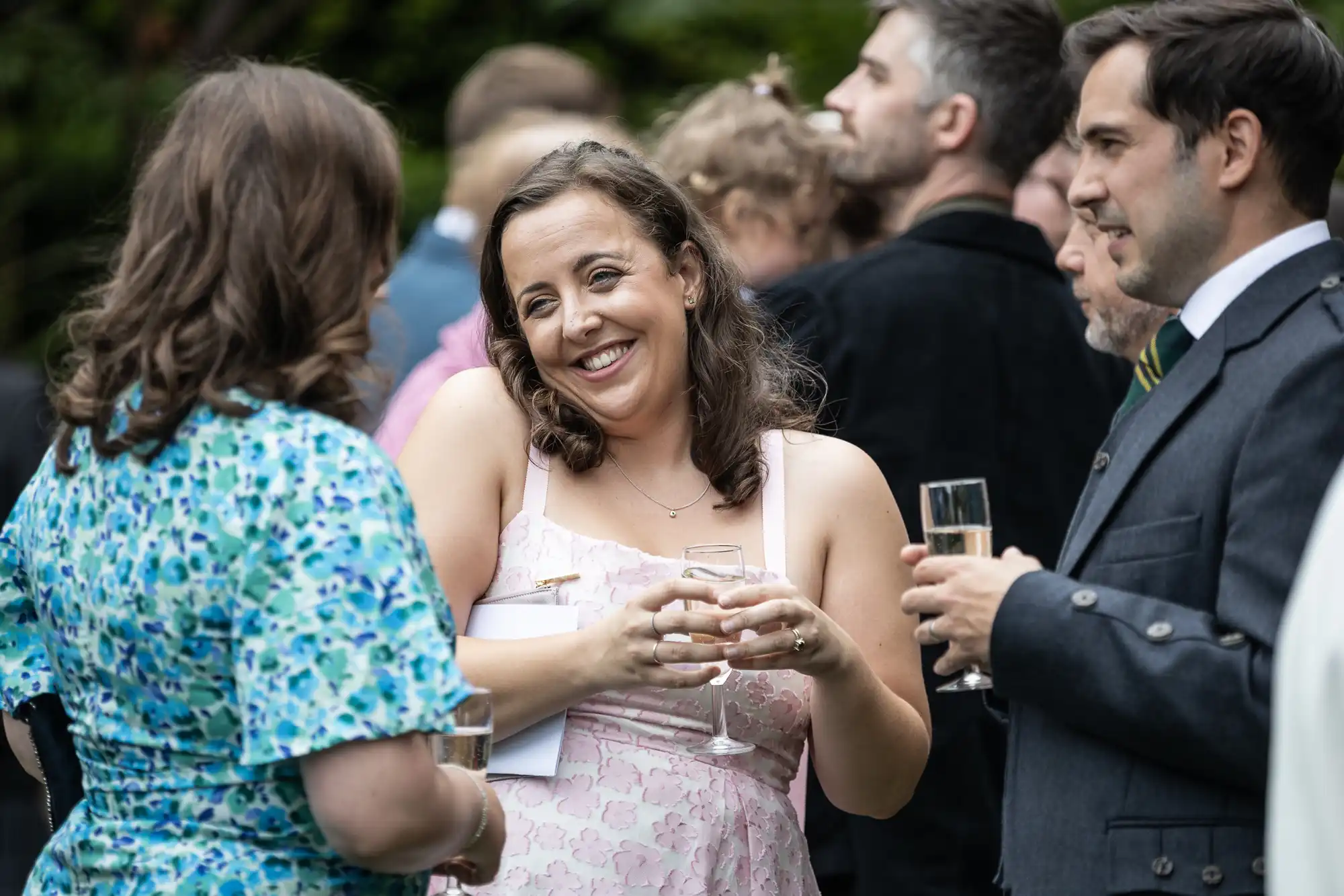 People at an outdoor social gathering, two women and one man in the foreground holding drinks, smiling and conversing.