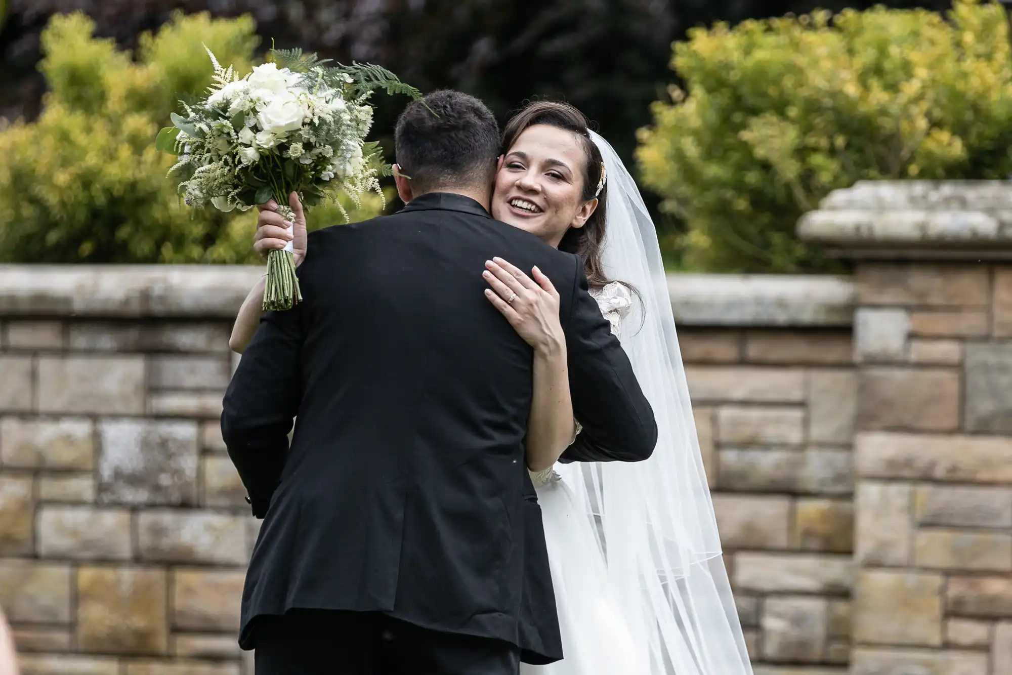 A bride in a white dress and veil hugs a groom in a black suit, smiling and holding a bouquet. They stand in front of a stone wall and greenery.