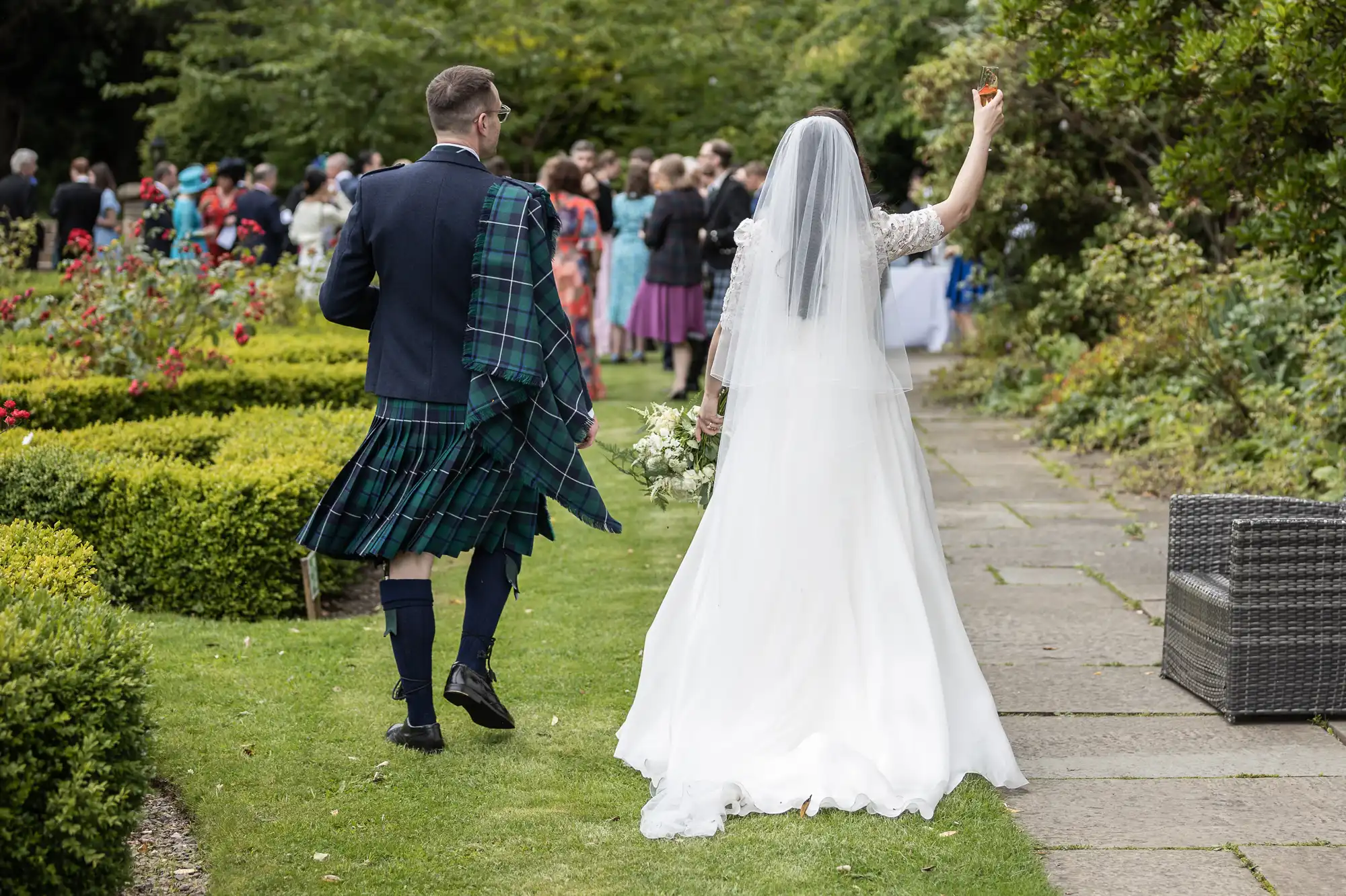 A bride and groom walk away on a garden path. The groom wears a kilt, and the bride holds a bouquet and raises a glass. People gather in the background.