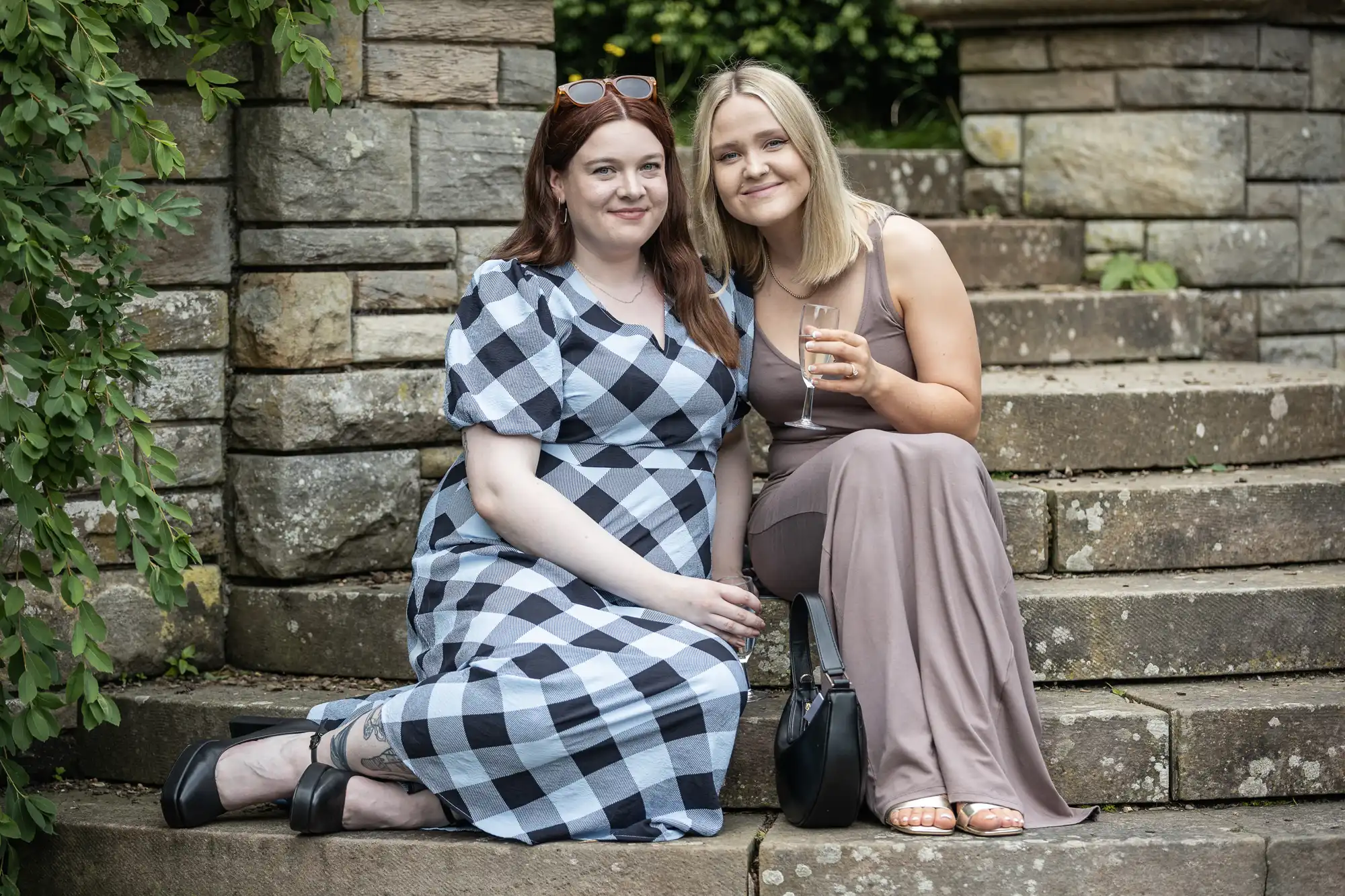 Two women seated on stone steps, one in a checked dress and the other in a beige dress, both smiling. The woman in beige holds a drink.