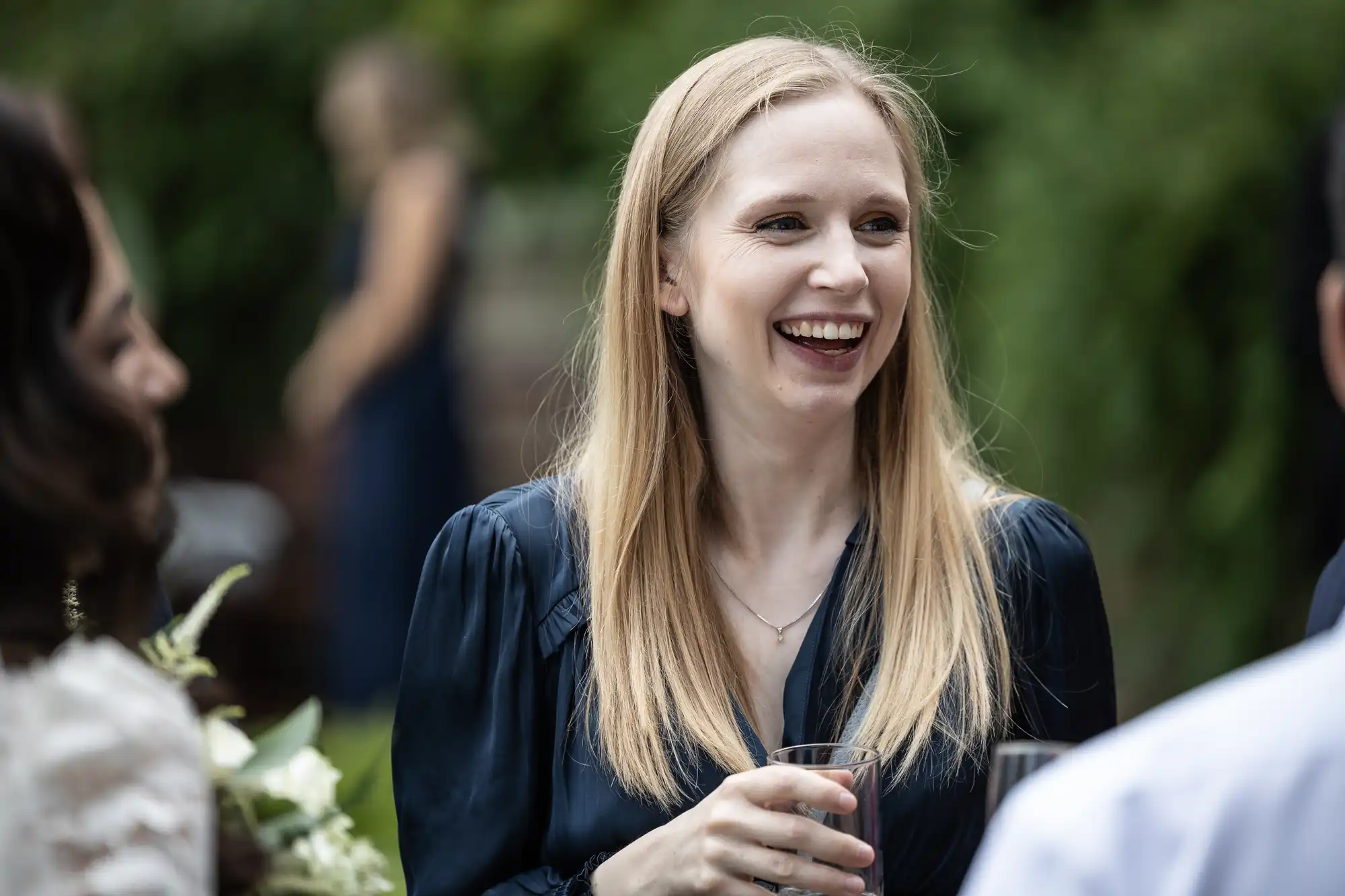 A woman with long blonde hair, wearing a dark top, is smiling while holding a glass. She is outdoors with greenery in the background.