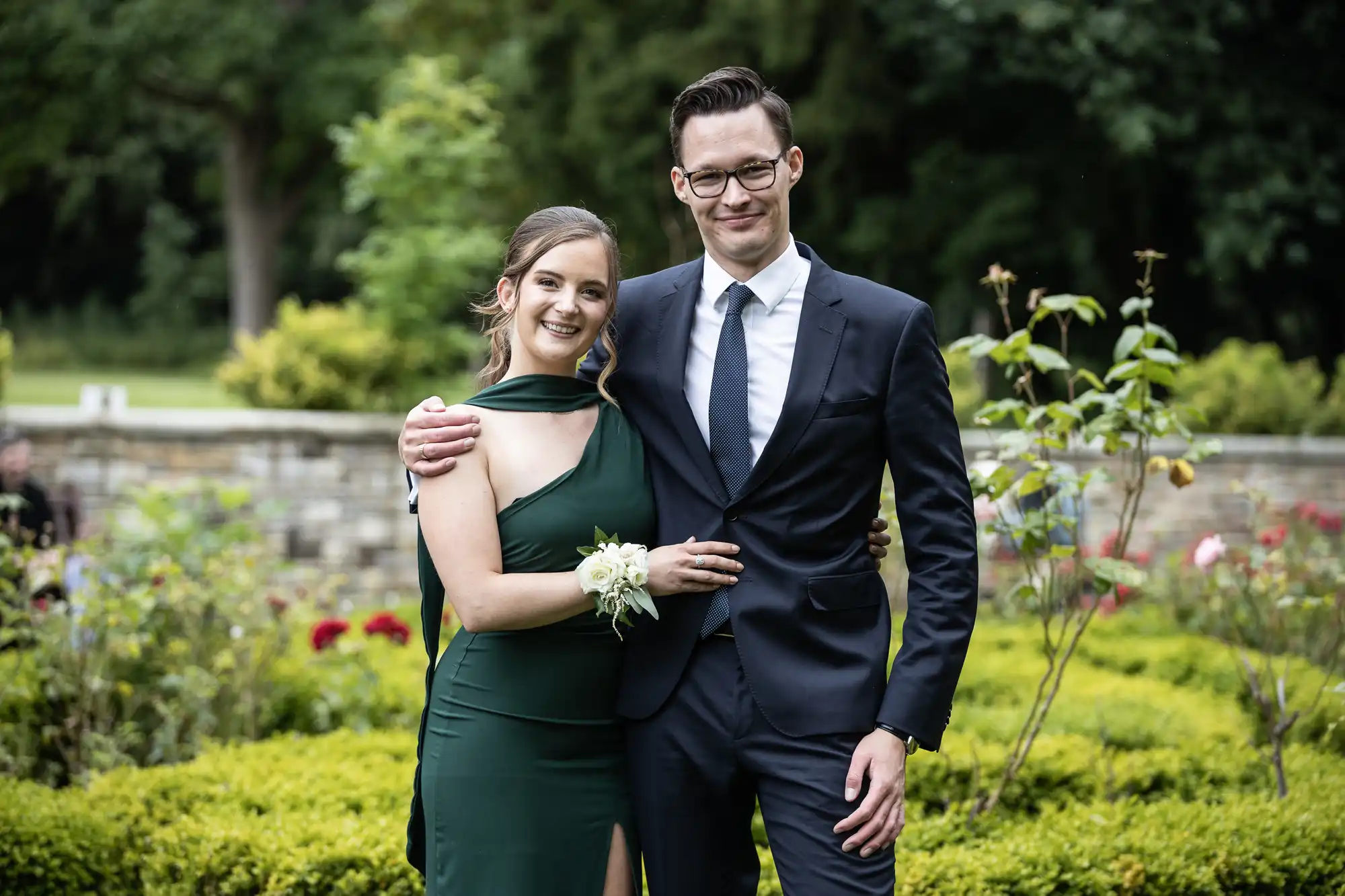 A woman and man stand together outdoors, smiling. She is in a green dress with a corsage, and he is in a dark suit and tie. They are in a garden with flowers and greenery.