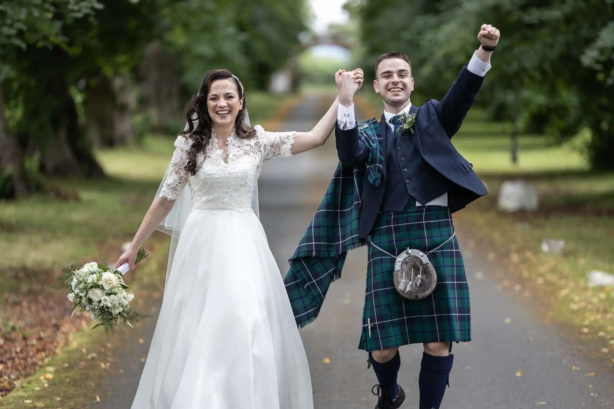 A bride and groom joyfully walk hand in hand. The groom wears a traditional kilt, and the bride holds a bouquet. They're outside on a tree-lined path.