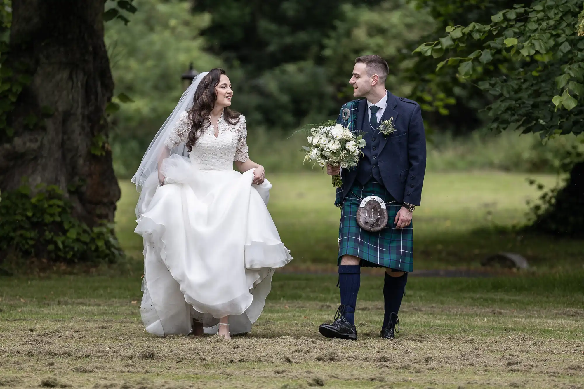 Bride and groom walking outdoors, the bride in a white dress and the groom in a kilt with a sporran, holding a bouquet.