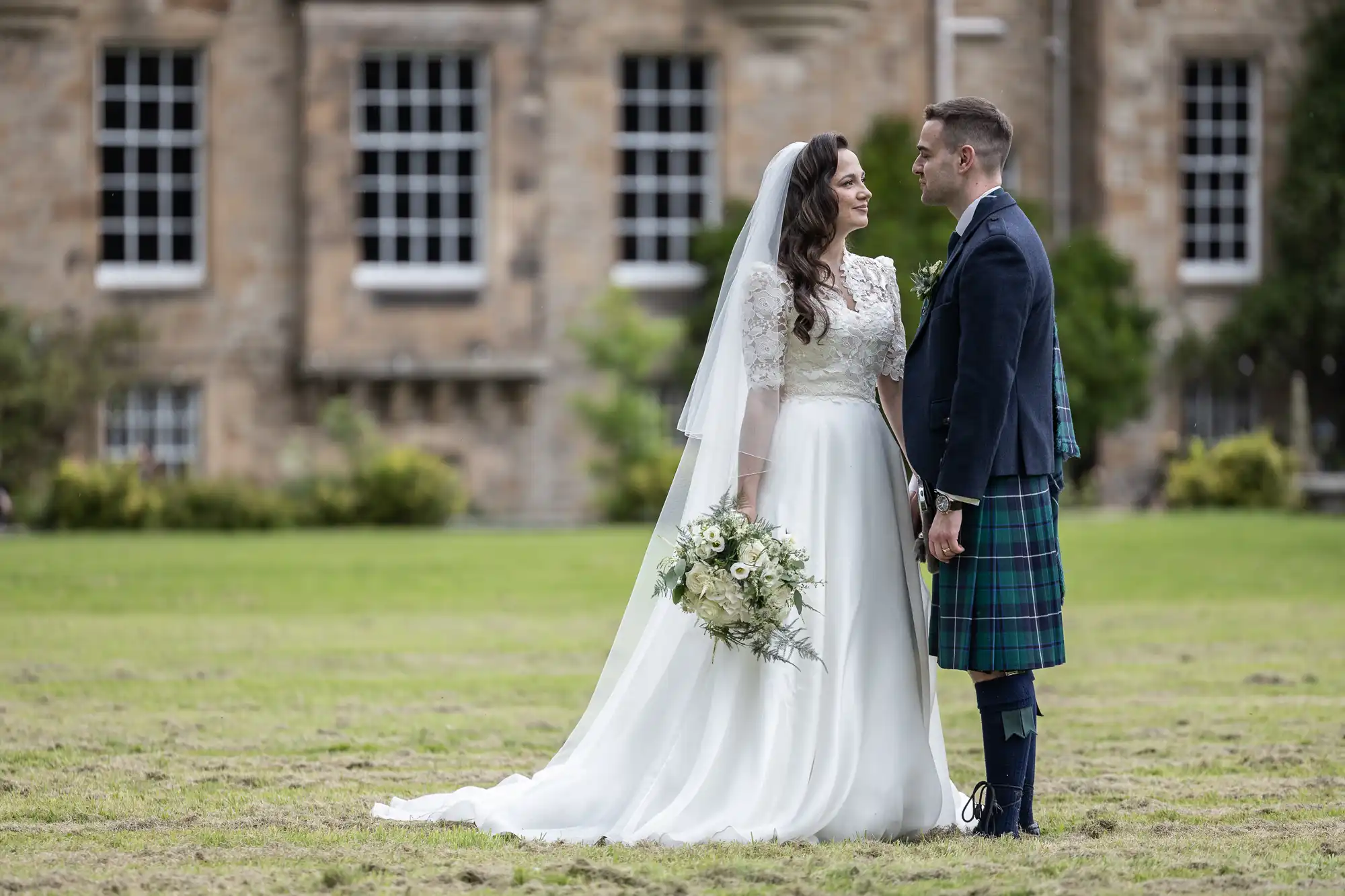 wedding photographer at Carberry Tower: Bride in white gown and groom in tartan kilt stand facing each other on grass, with brick building in the background.