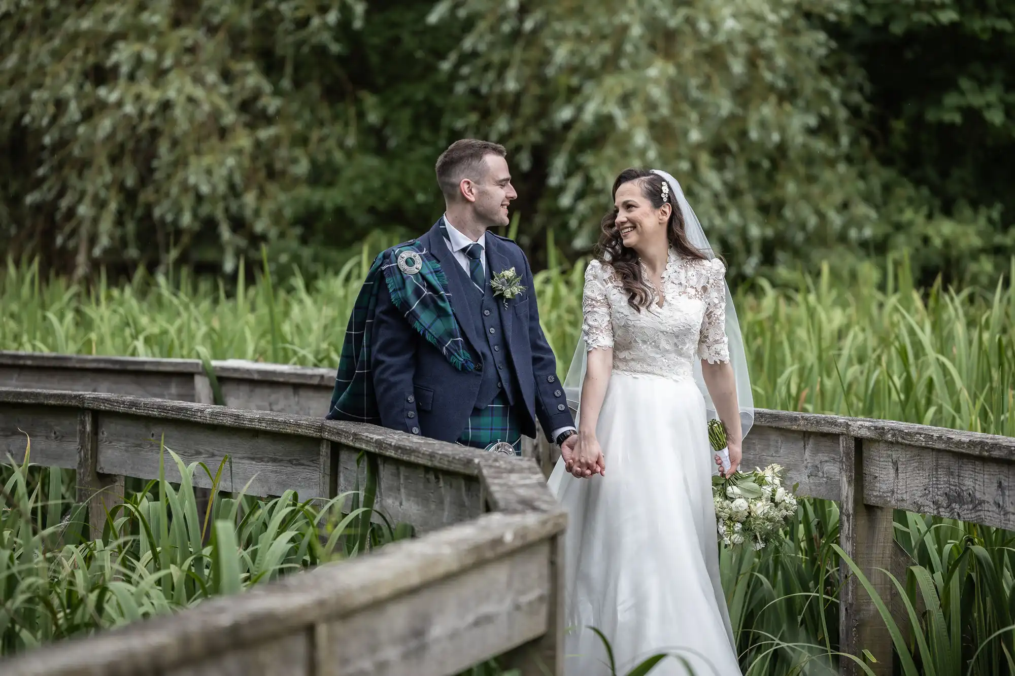 A bride and groom hold hands and smile at each other, walking on a wooden path surrounded by greenery.