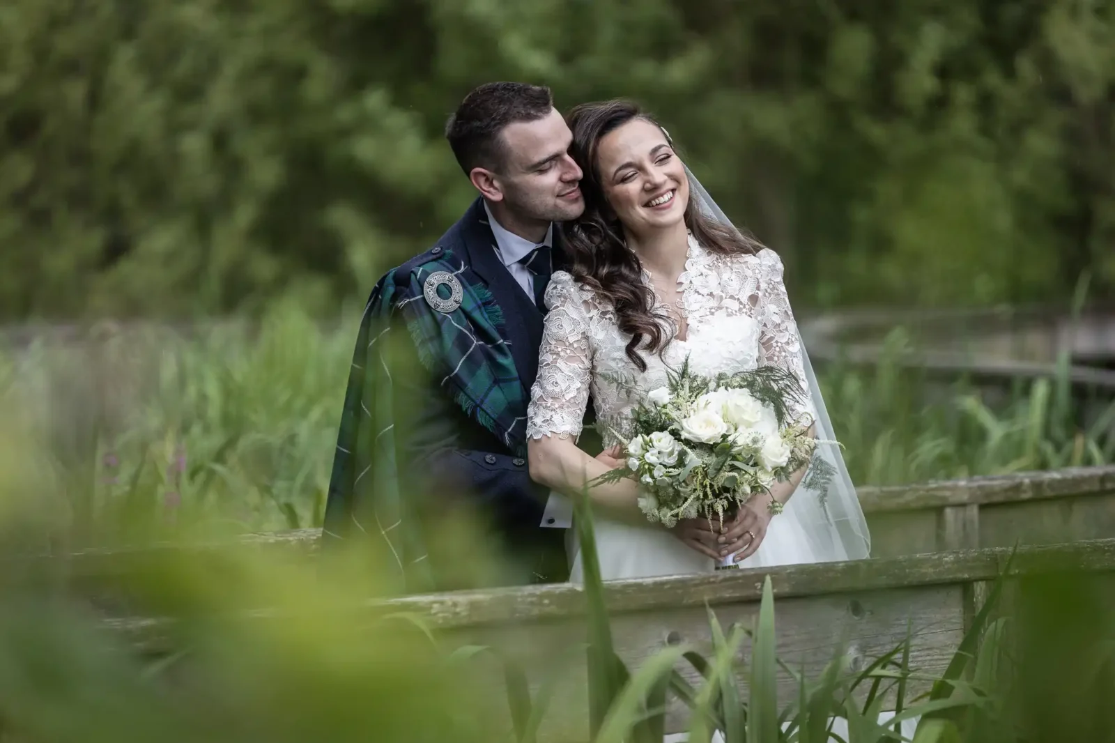 Carberry Tower wedding photographer image of a bride in a white dress holds a bouquet while a groom in a suit with a plaid shawl stands beside her. They are smiling outdoors, surrounded by greenery.