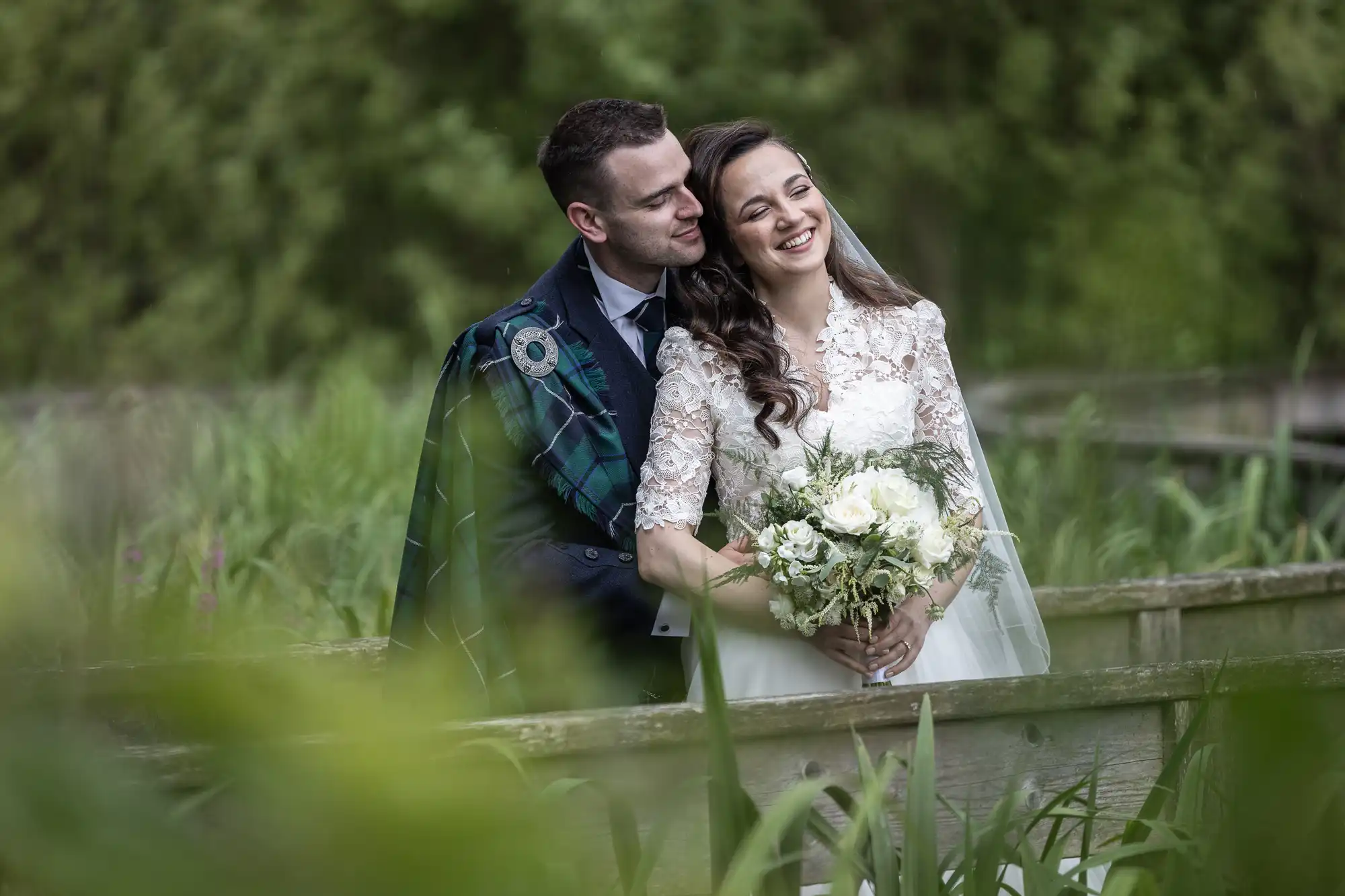 Carberry Tower wedding photographer image of a bride in a white dress holds a bouquet while a groom in a suit with a plaid shawl stands beside her. They are smiling outdoors, surrounded by greenery.