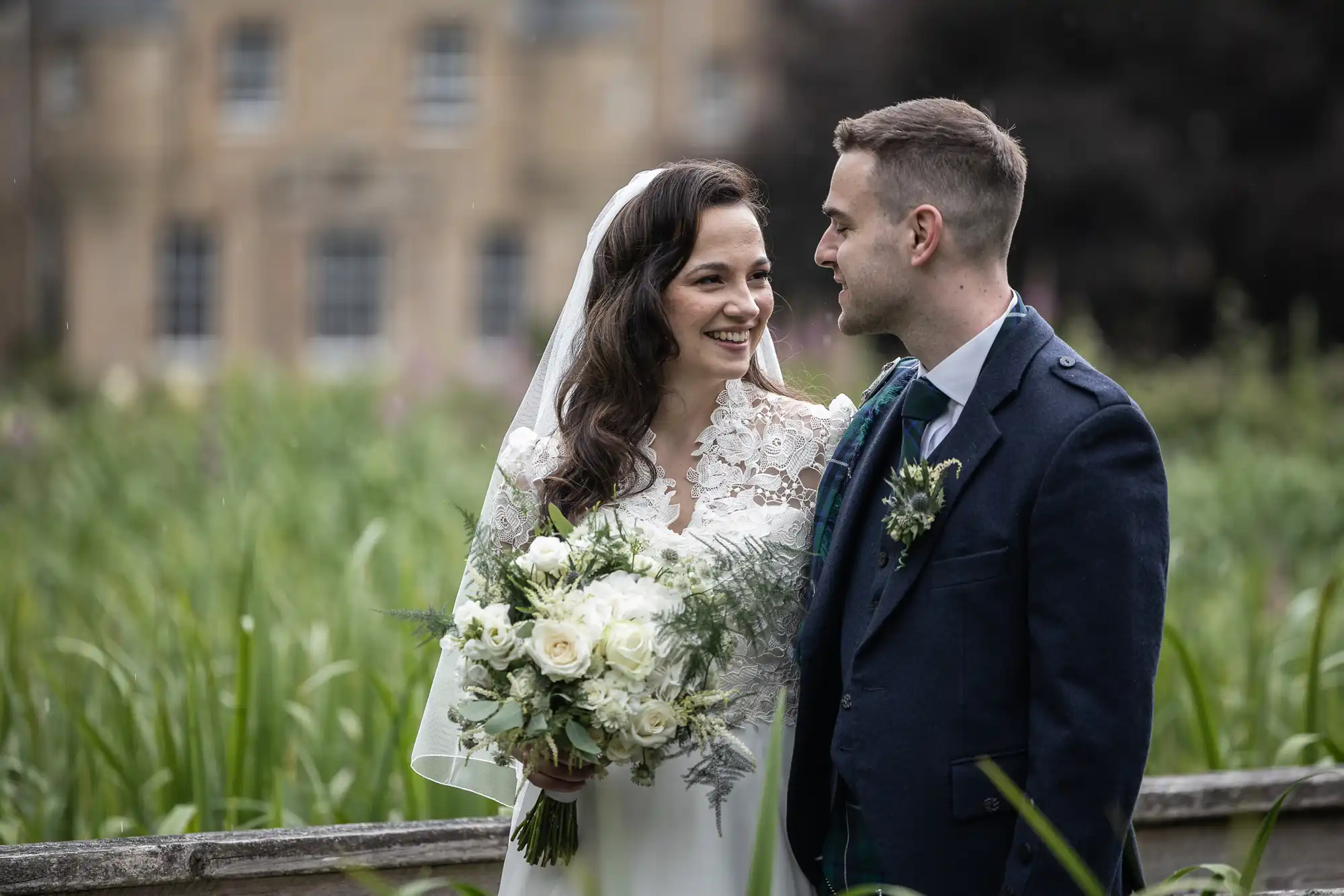 Bride and groom stand smiling at each other outside, with the bride holding a bouquet of white roses.