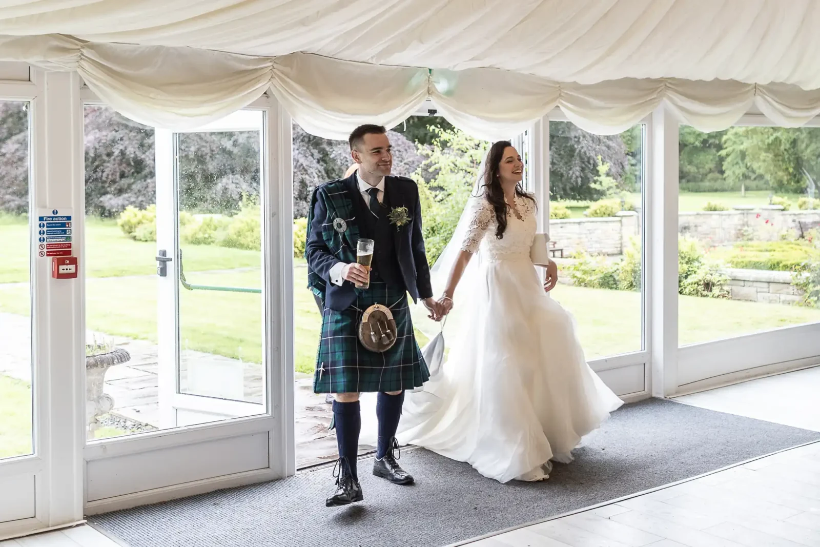 A bride and groom enter The Banqueting Pavilion at Carberry Tower holding hands. The groom wears a traditional Scottish kilt, and the bride wears a white gown.