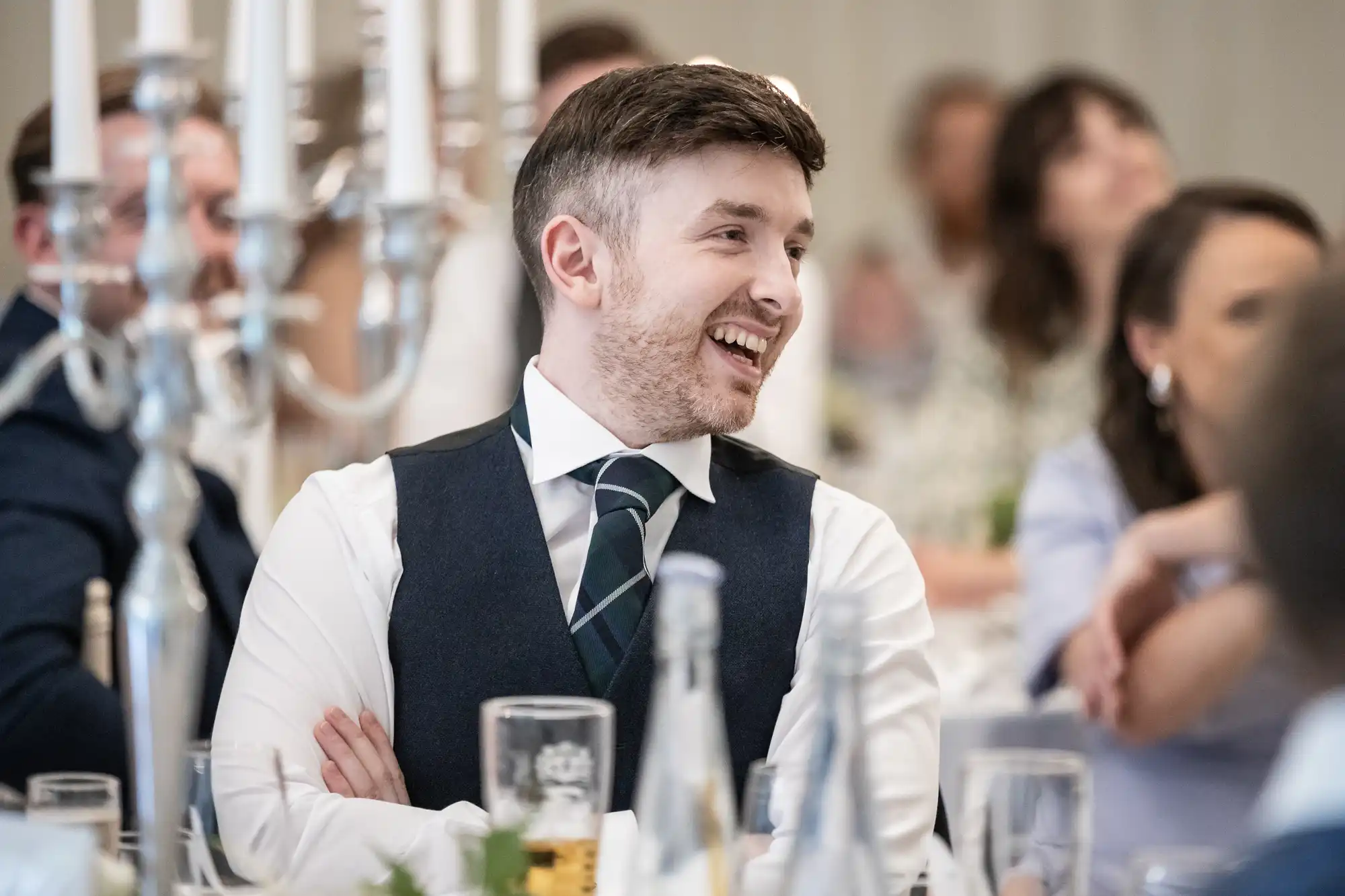Man in a vest and tie sitting at a table, smiling in a crowded room.