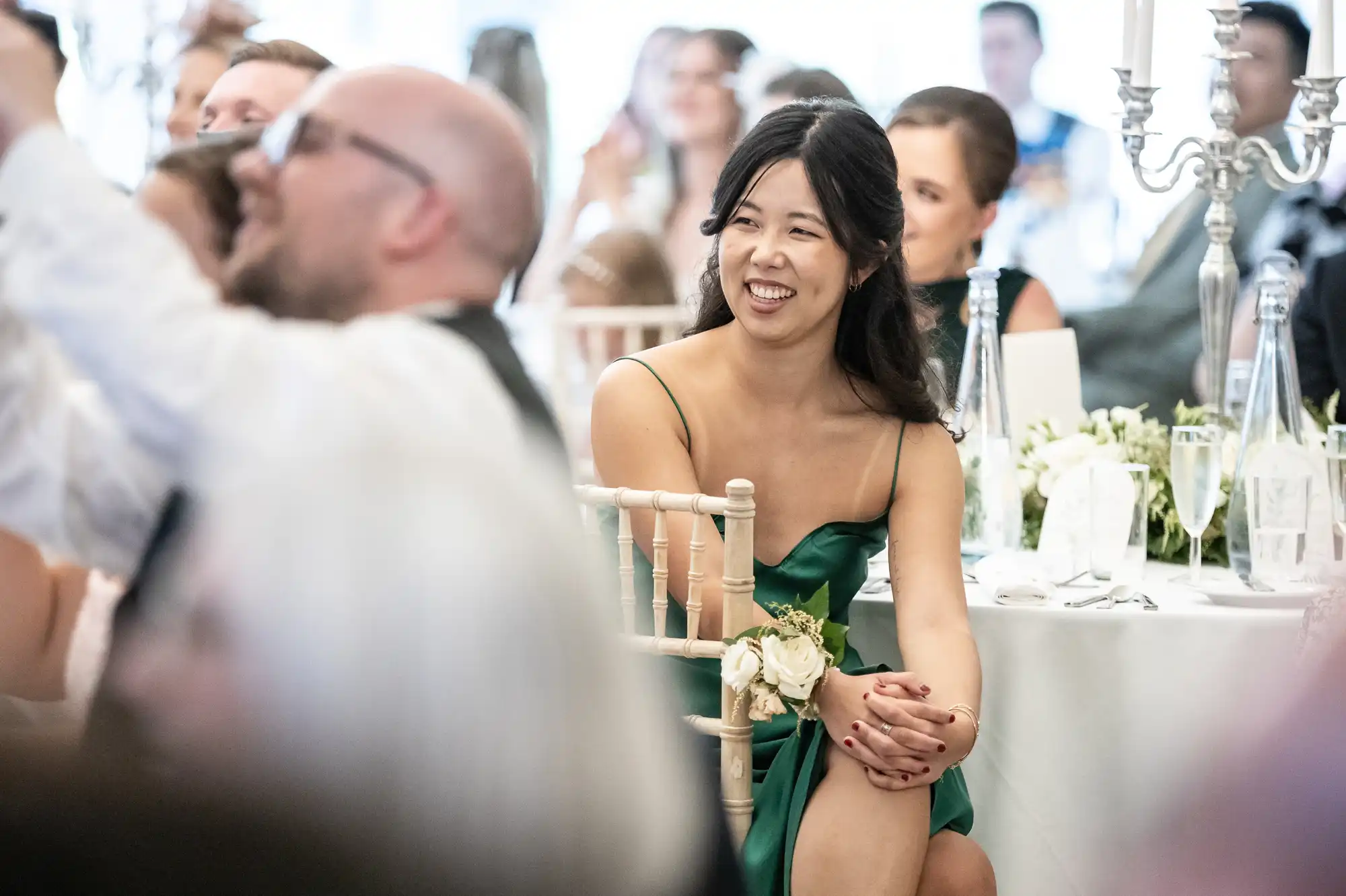 A woman in a green dress sits smiling at a formal event, surrounded by guests at elegantly set tables.