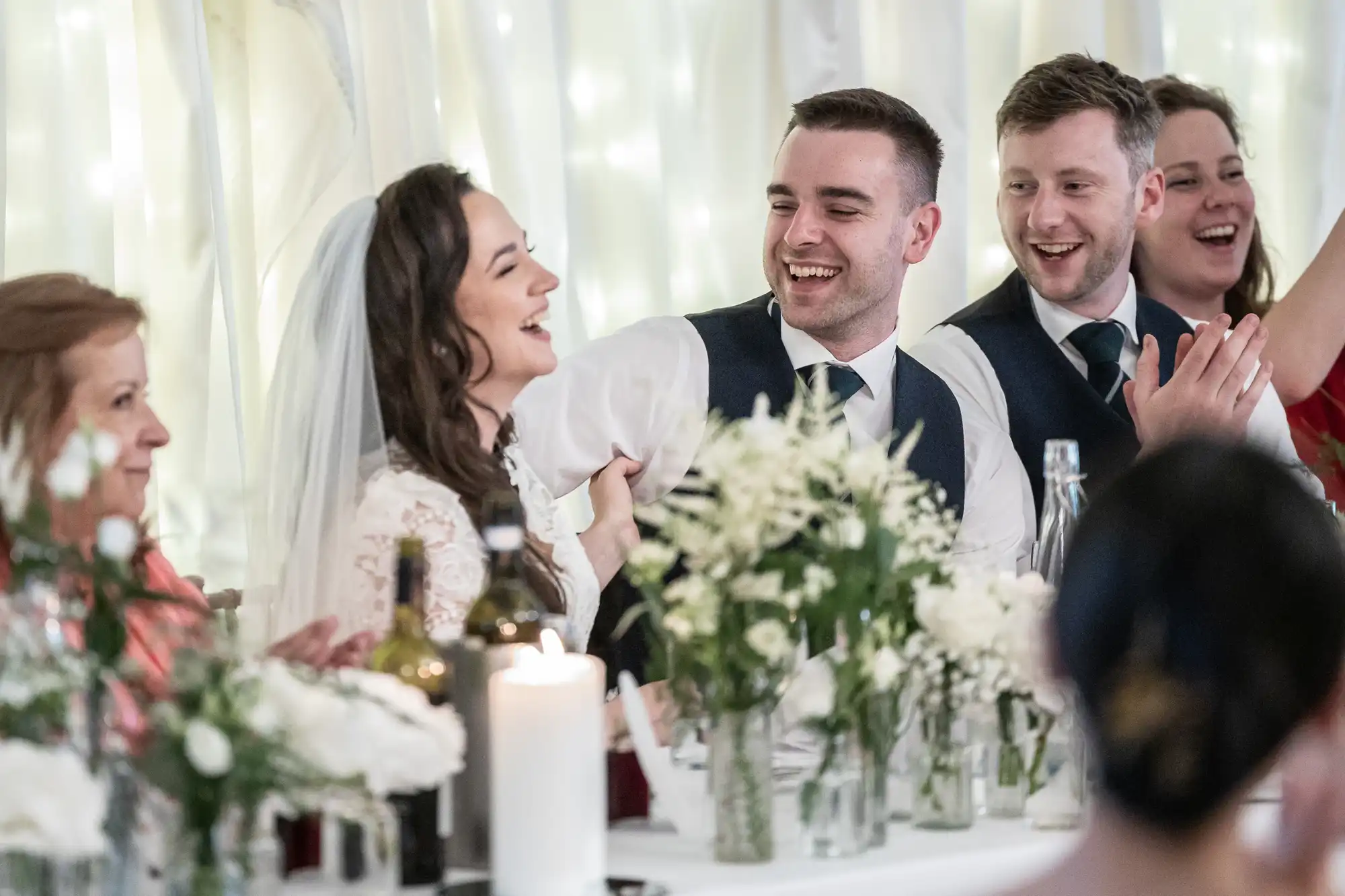 Bride and groom laughing at a wedding reception table with white floral decorations and other guests.