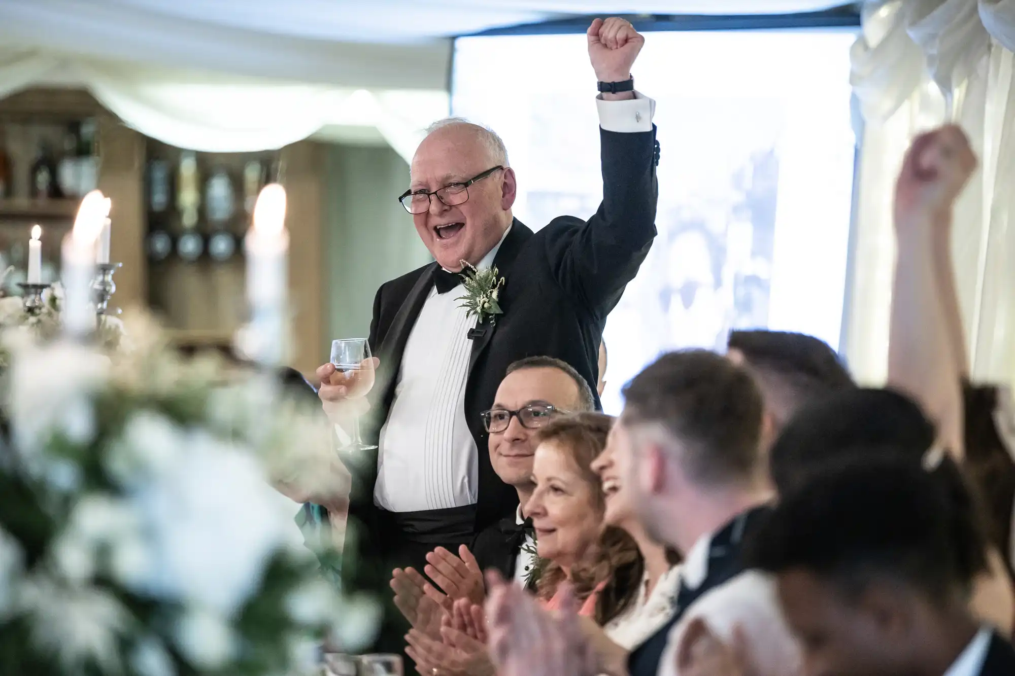 A man in a tuxedo raises his arm while holding a glass, smiling at a wedding reception. Guests around him are clapping and cheering.