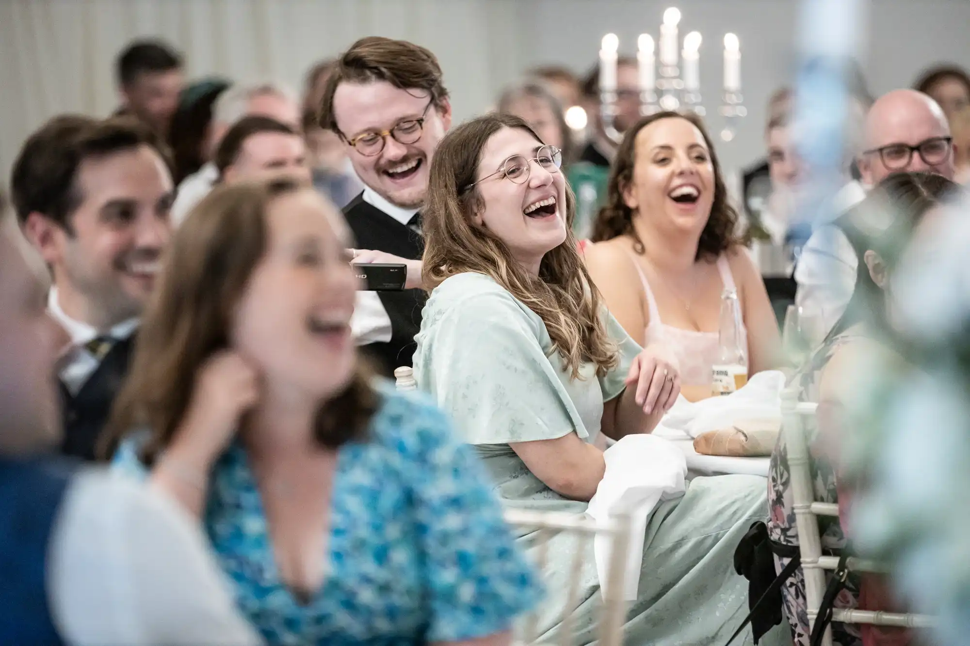 A group of people seated at tables, laughing and enjoying a social event. Candles are lit in the background.